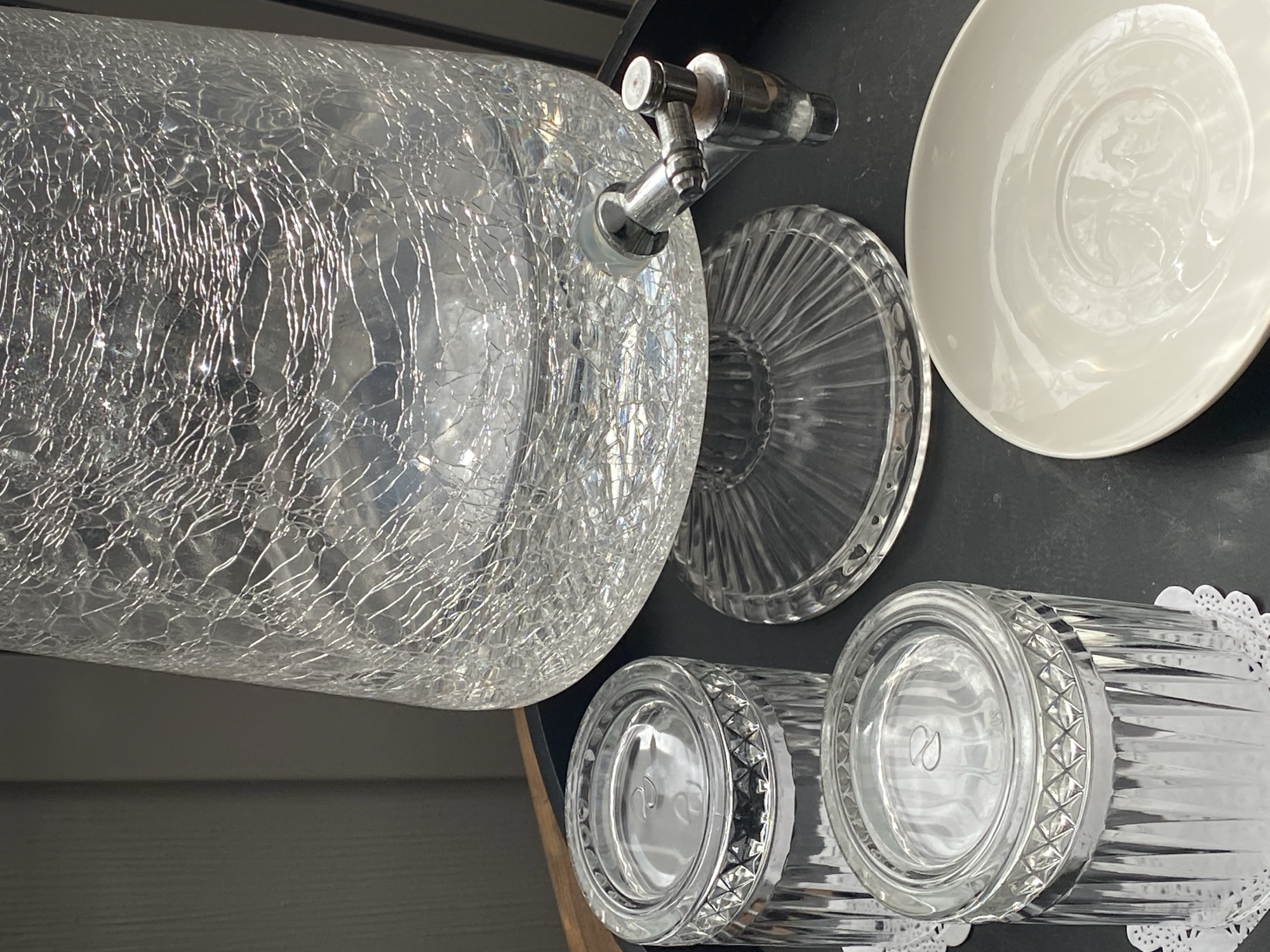 The image displays an indoor still life, focused on a beverage service setup presented on a dark, possibly black, rectangular tray.
A large, clear glass beverage dispenser occupies the right side of the tray. The dispenser features a distinctive "cracked ice" or craquelure texture and is filled approximately two-thirds with a clear liquid. A silver-toned spigot is attached to the lower right side of the dispenser. The dispenser rests on a separate, clear glass base with radiating fluted patterns.
To the left of the dispenser, two clear glass tumblers are positioned upside down. Both tumblers have vertically fluted sides and a decorative, possibly diamond-patterned, band near their rims (which are currently at the top). Each inverted tumbler rests on a small, white, circular paper doily. Visible on the bottom of the upper tumbler (now facing upwards) is a faint embossed "2".
In the lower right corner of the tray, a small, white, oval-shaped ceramic dish or shallow bowl is present.
The background appears out of focus, consisting primarily of a dark, uniform surface, with a lighter, possibly wooden, edge visible in the upper left background behind the tray. There are no identifiable visual markers within the image to indicate a specific location in the city of Votkinsk, Russia, as requested.