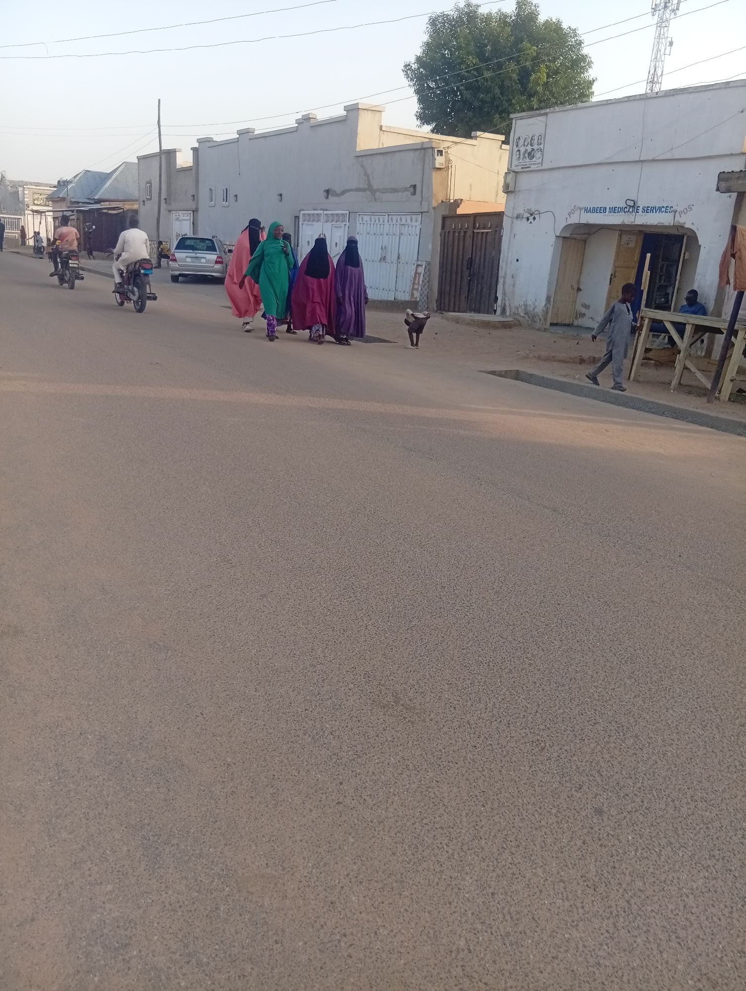 This image depicts a street scene in Dutse, Nigeria, likely in the late afternoon given the soft lighting. A group of five women, dressed in traditional Islamic attire including hijabs and long flowing robes in shades of pink, green, and purple, are walking together on the right side of the asphalt road. Two men on motorcycles are traveling in the same direction on the left side of the road, with one man in a light-colored traditional garment and the other in a darker outfit.
In the background, a row of low-rise buildings with light-colored facades lines the street. One building has signage that reads "HABEEB MEDICARE SERVICES". A young boy in a grey outfit is walking on the sandy area in front of the buildings, and further down, two men are seated on a wooden structure. A small black dog is visible in the foreground, near the women. The environment is dry and dusty, consistent with a Sahelian climate. The overall impression is of a typical urban street in a Nigerian city.
