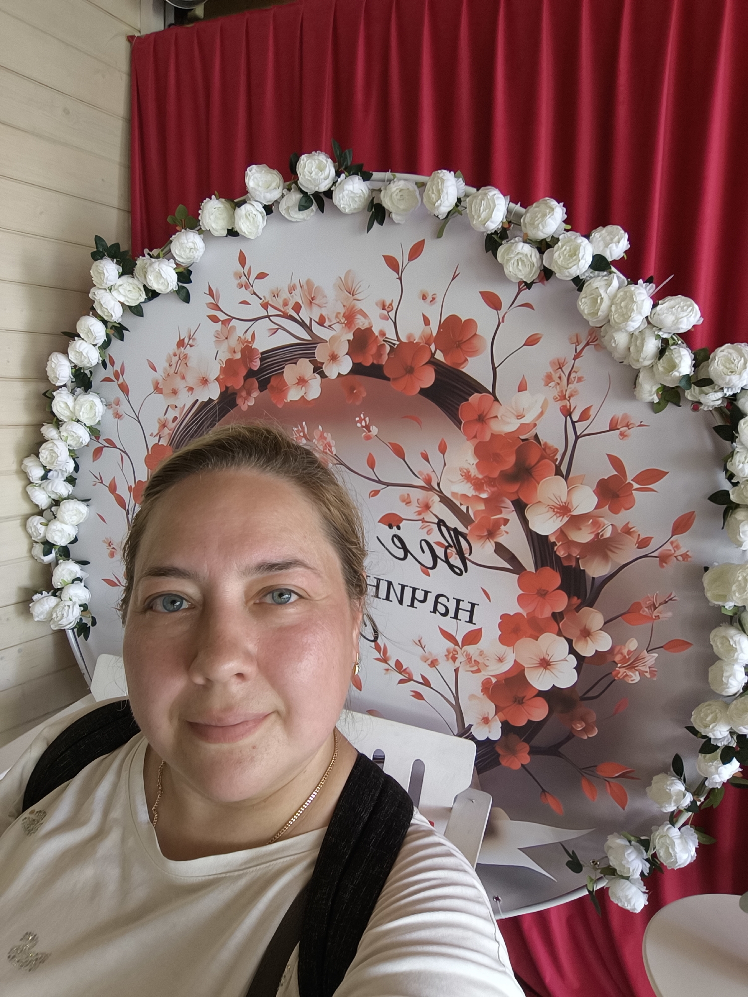 A woman with blonde hair and blue eyes is taking a selfie in front of a circular backdrop decorated with white and red flowers. The backdrop also features a floral design of branches with red and white flowers and leaves. Behind the backdrop, a red curtain is visible, and to the left, a light-colored wooden wall. The woman is wearing a white t-shirt with a gold chain necklace and a black backpack. The scene appears to be indoors, possibly at an event or photo booth, and the context of Taldom, Russia, suggests it might be a local celebration or gathering.
