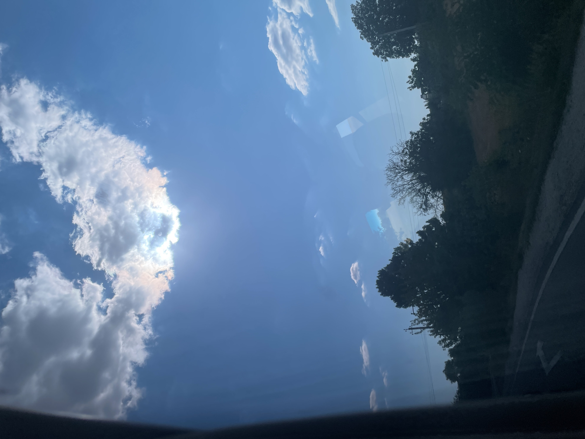 The image depicts a view of the sky and the landscape from inside a vehicle, likely on a road. The sky is a vibrant blue with scattered clouds, some of which are illuminated by the sun, creating a bright halo effect. The sun is partially obscured by a large, fluffy cloud, casting a warm glow. Below the sky, a treeline is visible, with various types of trees, some dense and leafy, others bare. A road with a white arrow marking can be seen in the foreground, suggesting a journey or travel. The overall impression is of a sunny day in a rural or suburban setting.