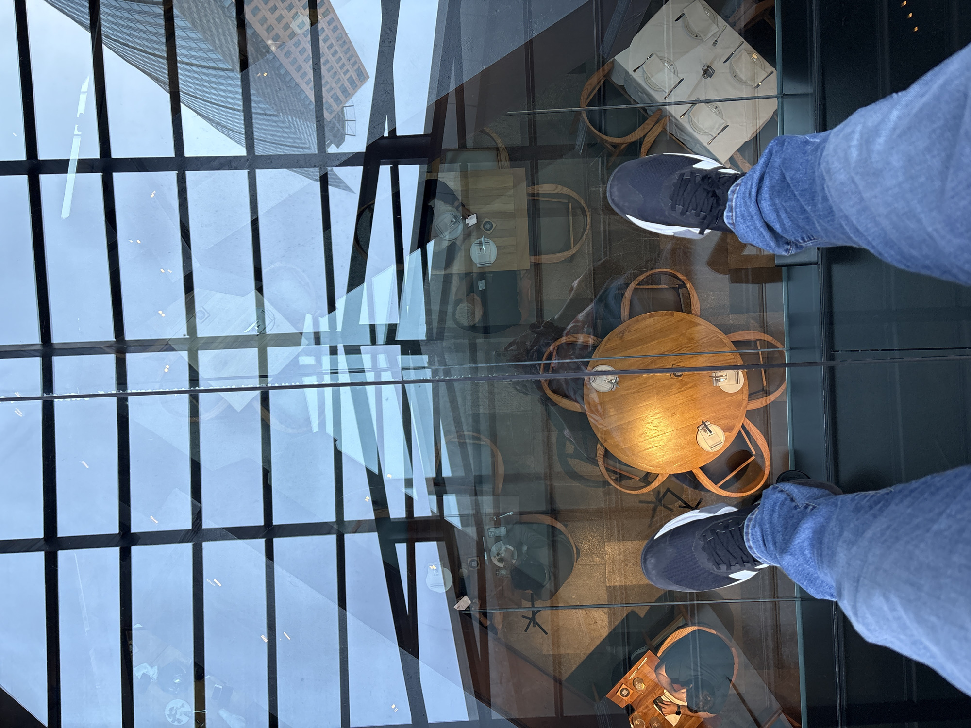 The image displays a first-person, downward perspective of an individual's lower legs and feet, clad in blue jeans and dark sneakers with white accents, standing on a clear glass floor. Directly beneath the transparent floor, a sophisticated restaurant or cafe environment is visible. This space contains multiple circular wooden tables surrounded by chairs, some featuring dark upholstery and others with wooden frames. Several tables are set with white plates, cutlery, and glassware, indicating active service. Reflected on the glass surface are elements of the interior ceiling, which consists of a grid-patterned glass structure, and a prominent modern skyscraper exterior characterized by an angular, grid-like facade, consistent with the Torre Reforma building. The location is a high-level observation point, likely a sky bar or terrace such as the Cityzen Rooftop Bar at the Hotel Sofitel Mexico City Reforma, overlooking an internal dining area and offering reflections of the urban landscape in Mexico City, Mexico.