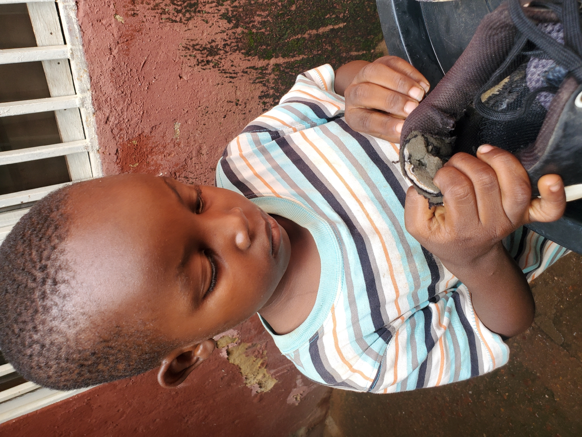 This media file features a close-up, high-angle shot of a young Black child wearing a horizontally striped t-shirt in shades of blue, orange, and white. The child has their eyes closed, appearing to be concentrating or resting. They are holding a black shoe with a torn sole, with their fingers manipulating the damaged area of the shoe. The background consists of a textured, reddish-brown wall with some patches of green moss or algae. To the left of the child, there is a white metal grate or window bars. The ground appears to be wet and possibly muddy or paved, with some debris scattered on it. The lighting suggests it could be daytime, possibly overcast or shaded. The overall scene conveys a moment of interaction with a worn-out object, possibly indicating poverty or resourcefulness. The child's expression is calm, without any discernible strong emotion. No visible text is present in the image. The location context is Jos, Nigeria.