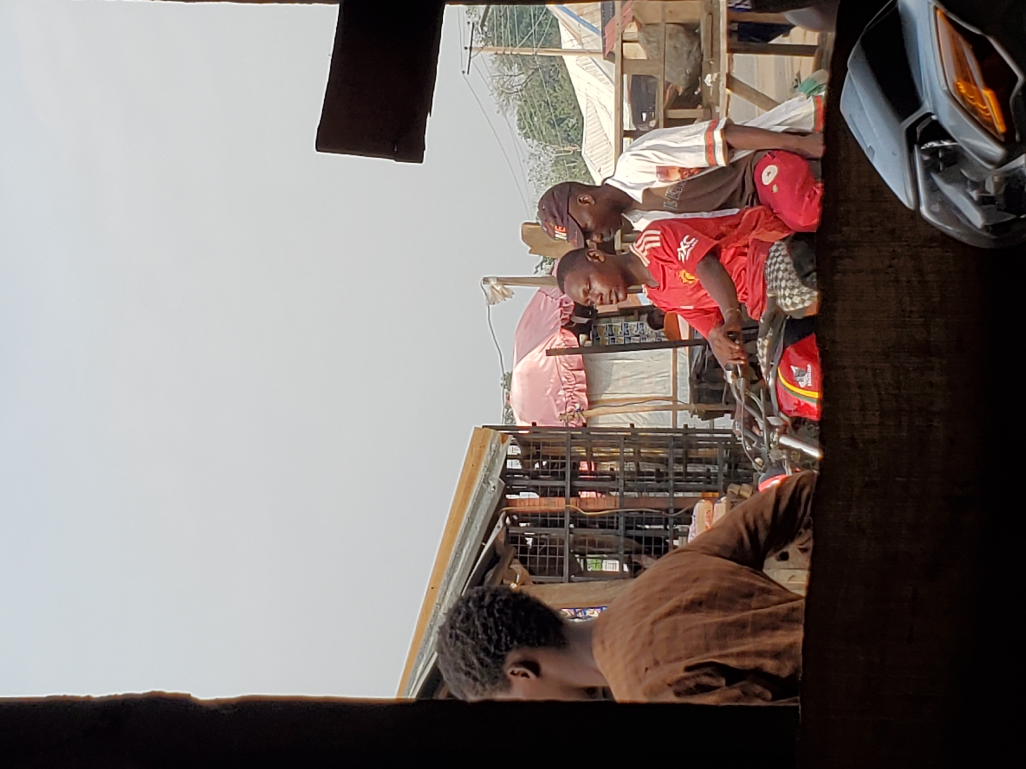 The image shows a street scene in Mararaba, Nigeria, viewed from an elevated position. Two men are sitting on a motorcycle, one wearing a red Manchester United jersey. A third man, in a brown shirt, is visible in the foreground, likely observing the scene. The background reveals a bustling street with shops and other commercial activity, including umbrellas and various metal structures. A car's front end is visible on the right. Overhead power lines and other urban infrastructure elements are present.