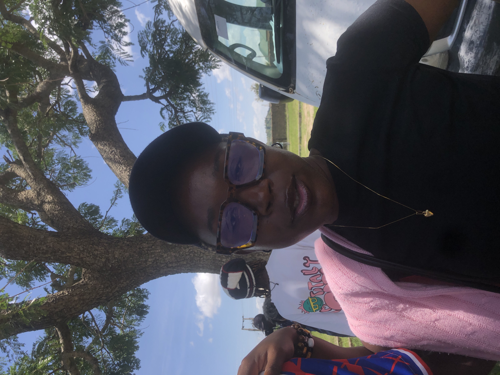 A medium shot captures a Black woman in the foreground, facing the camera, wearing a black t-shirt, a pink knitted cardigan, a black baseball cap, and tortoiseshell glasses. She has a gold locket on a thin chain around her neck. To her left, another person is partially visible, wearing a colorful patterned shirt and bracelets. In the background, a white van is parked to the right of the frame. Behind the woman and to the left, another individual wears a white t-shirt with a graphic design and a black and white patterned beanie. The scene is outdoors, under a large tree with green leaves, with a blue sky and scattered clouds overhead. The ground appears to be grassy and uneven. The overall impression is of a casual outdoor gathering or event. The location is identified as Gwafan, Nigeria.