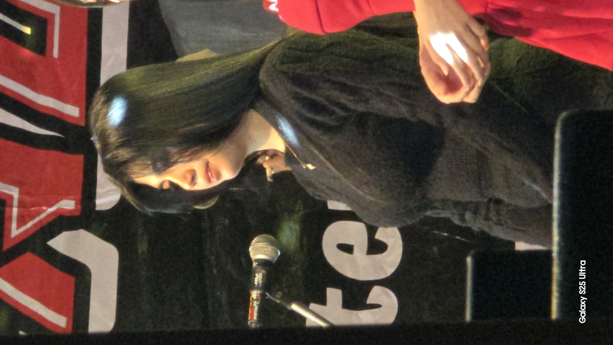 A young woman with long, dark hair stands on a stage in front of a banner with red and white lettering. She is wearing a black, long-sleeved, textured sweater and a necklace with a heart-shaped pendant. A microphone is visible to her left, and a portion of another person in a red dress is to her right. The word "te" is printed in large white letters on a dark background behind her. The text "Galaxy S25 Ultra" is visible in the lower left corner of the image. The image appears to have been taken during a performance or event.