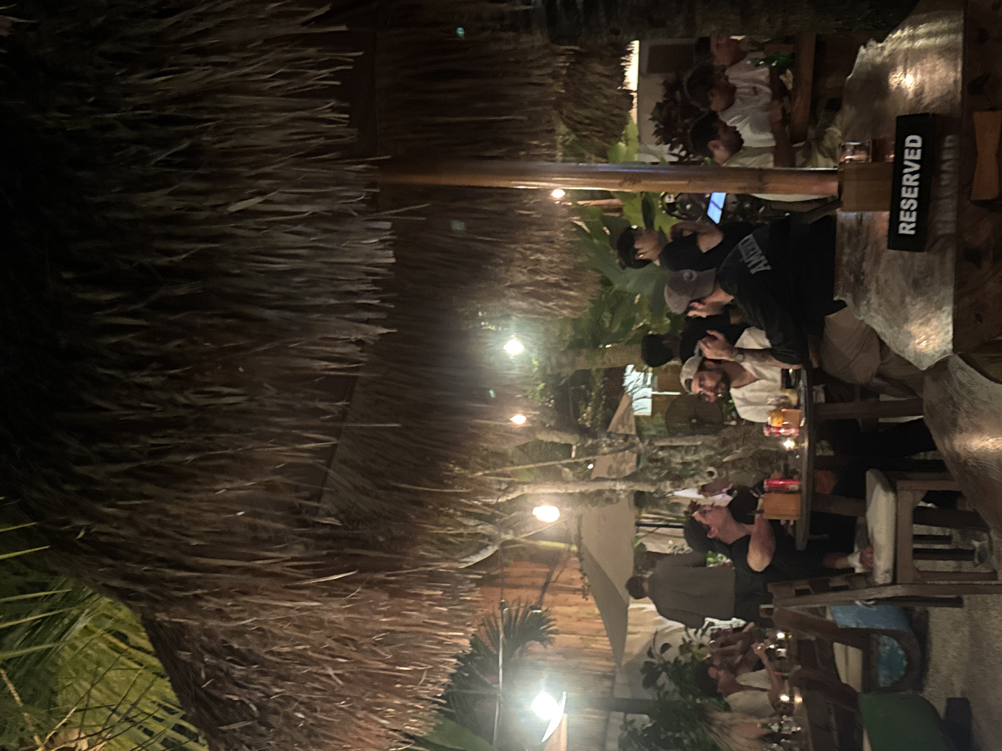 A sideways, medium-shot photograph shows an outdoor dining area in Kuta Selatan, Indonesia. A thatched roof covers an area where people sit at tables. A black sign on the upper right table reads "RESERVED." People are sitting and standing at the tables. Some appear to be talking and eating. The area is illuminated by lights. Palm trees are visible in the background.