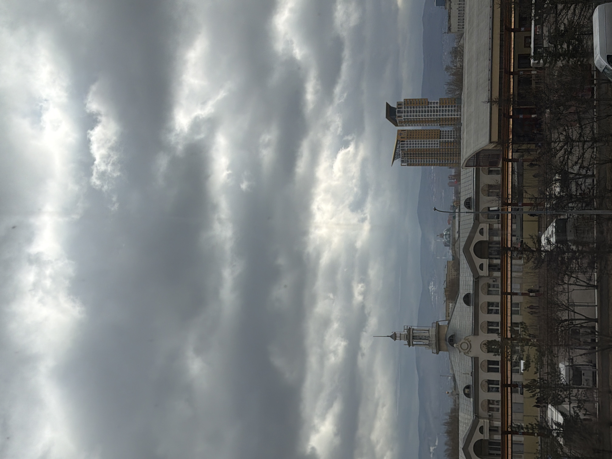 The image captures an urban landscape viewed from an eye-level perspective under a heavily clouded sky. The photograph is oriented vertically, but when rotated to landscape, it reveals a city scene stretching towards distant mountains.
The sky is a prominent feature, filled with dense, grey clouds, but bright patches indicate sunlight breaking through, creating visible shafts of light. In the mid-ground, on the left side of the frame, two modern high-rise buildings stand out. They feature numerous windows and distinct yellow-gold architectural panels, topped with dark, angular roofs. These structures are positioned against a backdrop of hazy, grey mountains or hills that define the distant horizon.
Closer to the foreground, on the right, is a more classically styled multi-story building. It has an ornate facade with multiple arched windows and is distinguished by a prominent turret or spire on its roof. Bare-branched trees are visible in front of this building, suggesting a colder season like late autumn or winter. Several light-colored vehicles, including vans, are partially visible on what appears to be a street or parking area near the building.
The scene depicts a static city environment during daytime, likely late morning or afternoon, given the quality of light. The weather is overcast but with moments of brightness. No specific human activity is evident, and no discernible text is visible anywhere in the image. The blend of modern and traditional architecture with a mountainous backdrop is characteristic of a city like Krasnoyarsk.