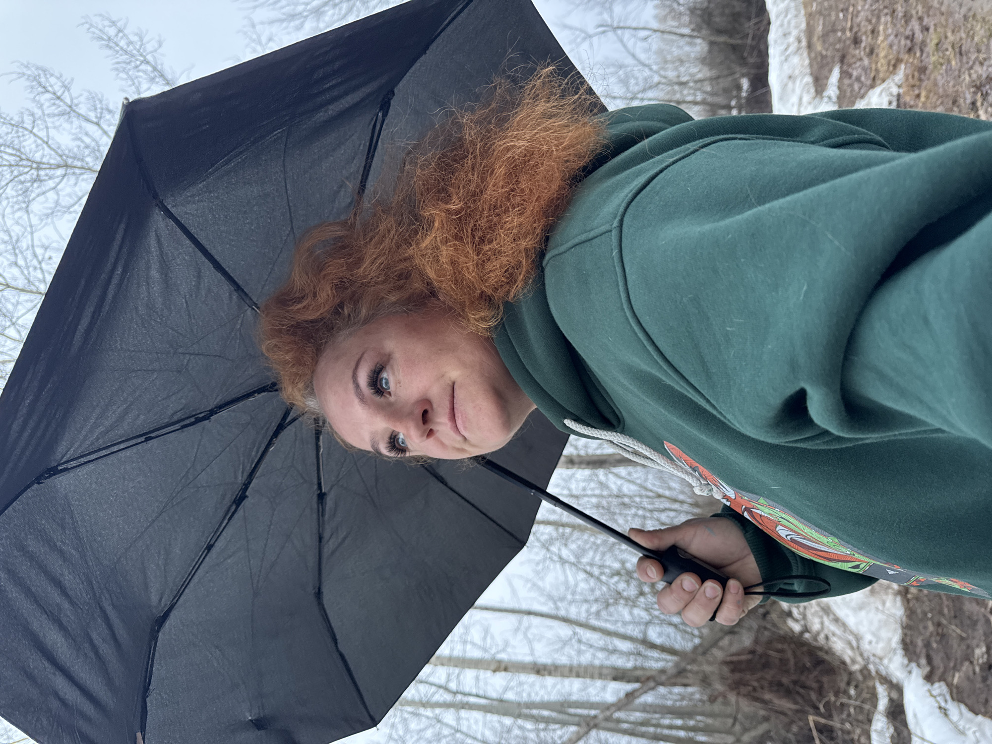 A woman with red curly hair and blue eyes is holding a black umbrella. She is wearing a green hoodie with a graphic design on the sleeve. The background shows a snowy landscape with trees, suggesting a winter scene. The exact location in Chaikovsky, Russia is not evident from the image.