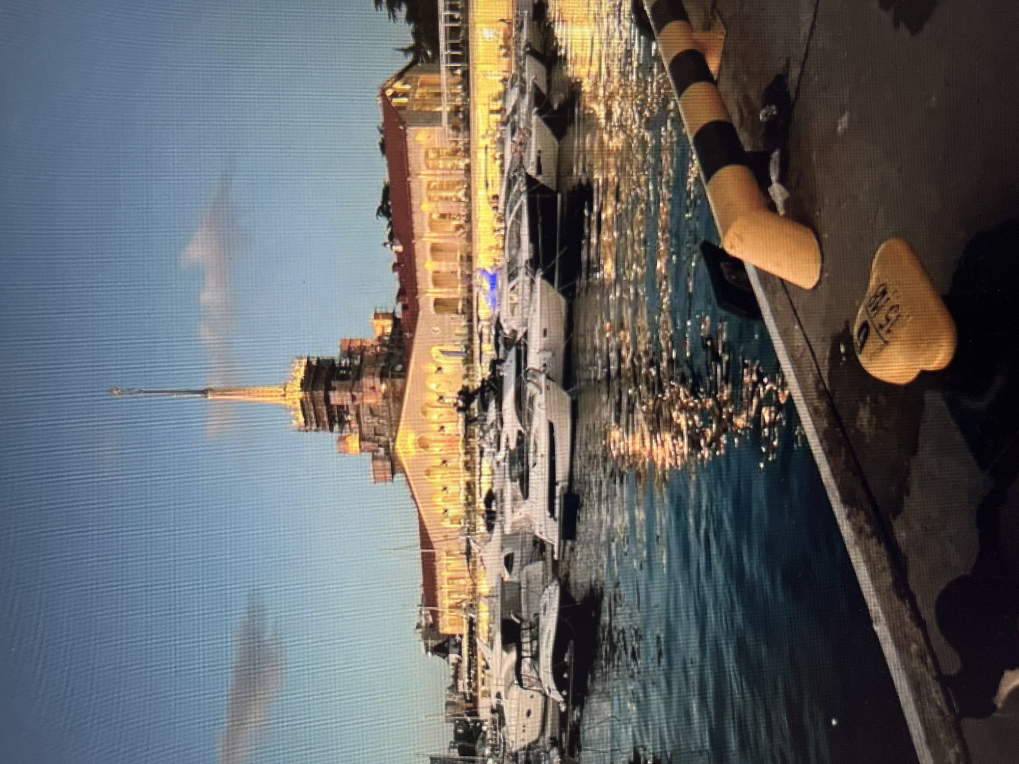 A vertical, medium shot captures a marina at dusk with a prominent building in the background. The building features a tall, spire-like tower at its center, illuminated with yellow lights. The main structure of the building is a long, two-story edifice with a red-tiled roof and numerous arched windows, also lit from within. The sky above is a pale blue with a few wispy clouds.
In the foreground, several white yachts are docked in the dark blue water. Their reflections shimmer on the water's surface, along with the lights from the building. The water itself appears calm, with gentle ripples.
To the right of the frame, a concrete pier or walkway is visible. On this surface, a yellow and black striped bollard or barrier extends towards the water, and a yellow, mushroom-shaped buoy with black markings, possibly a number "15 MB," rests on the ground. The overall lighting suggests twilight or early evening. The location is identified as the Sea Terminal in Sochi, Russia.