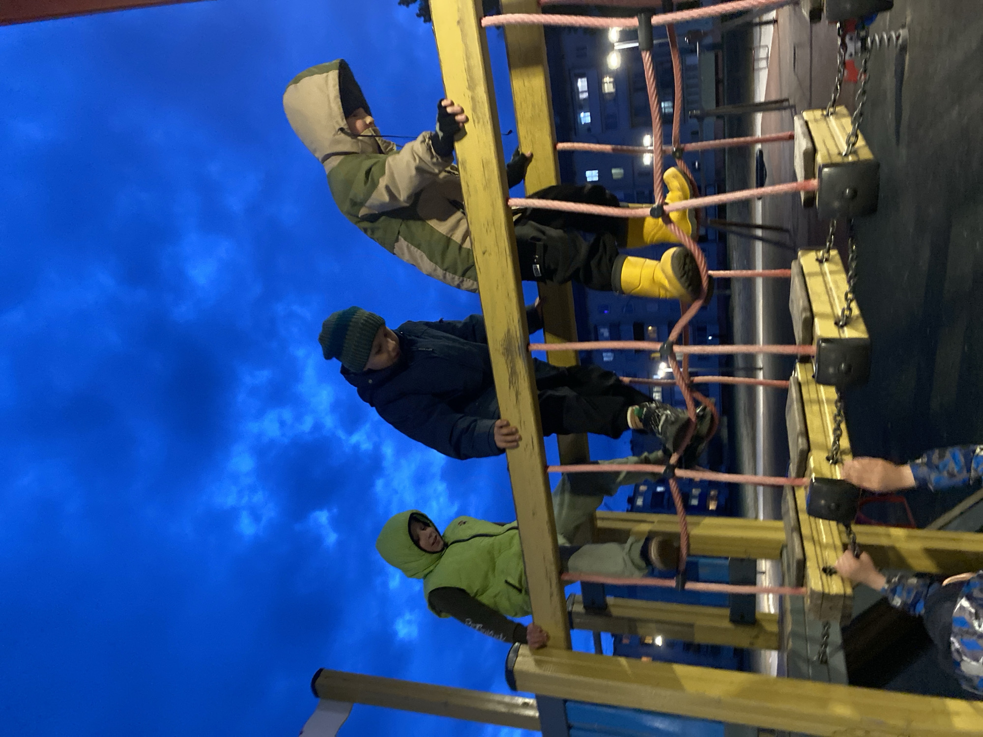 Three children are on a playground climbing structure made of yellow wooden supports and pink rope netting. The child at the bottom is wearing a light green vest. The child in the middle is wearing a dark blue jacket and a dark blue beanie. The child at the top is wearing a camouflage jacket and yellow rubber boots. The sky is dark blue, indicating it is evening or night. In the background are windows of an apartment building, which is in Noviy, Russia. Two hands are holding onto the side of the playground equipment on the right.