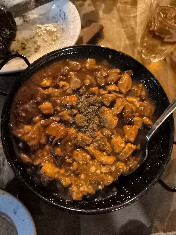 This close-up image depicts a rustic meal served in a warm, dimly lit setting, likely a restaurant or tavern in Nea Erithrea, Greece, during the evening. The main subject is a black, round pan, possibly cast iron or enamel, brimming with a hearty stew. The stew consists of numerous cubed pieces of meat (likely pork or beef) immersed in a rich, reddish-brown sauce. A generous topping of dried herbs, resembling oregano, is sprinkled over the center of the dish. A silver spoon is partially visible within the stew, indicating it is either being served or actively consumed.
Surrounding the main dish, a used white plate with remnants of sauce and food particles is visible on the top left, suggesting a meal in progress. In the upper right corner, a glass holds a golden-brown liquid, possibly a spirit like whiskey or another beverage. The table surface appears to be a warm, light brown, potentially wood or a textured tablecloth. A small, partially visible blue and white ceramic or stone object is present in the bottom left. The overall ambiance is cozy and intimate, enhanced by the artificial, warm lighting. There is no visible text in the image.