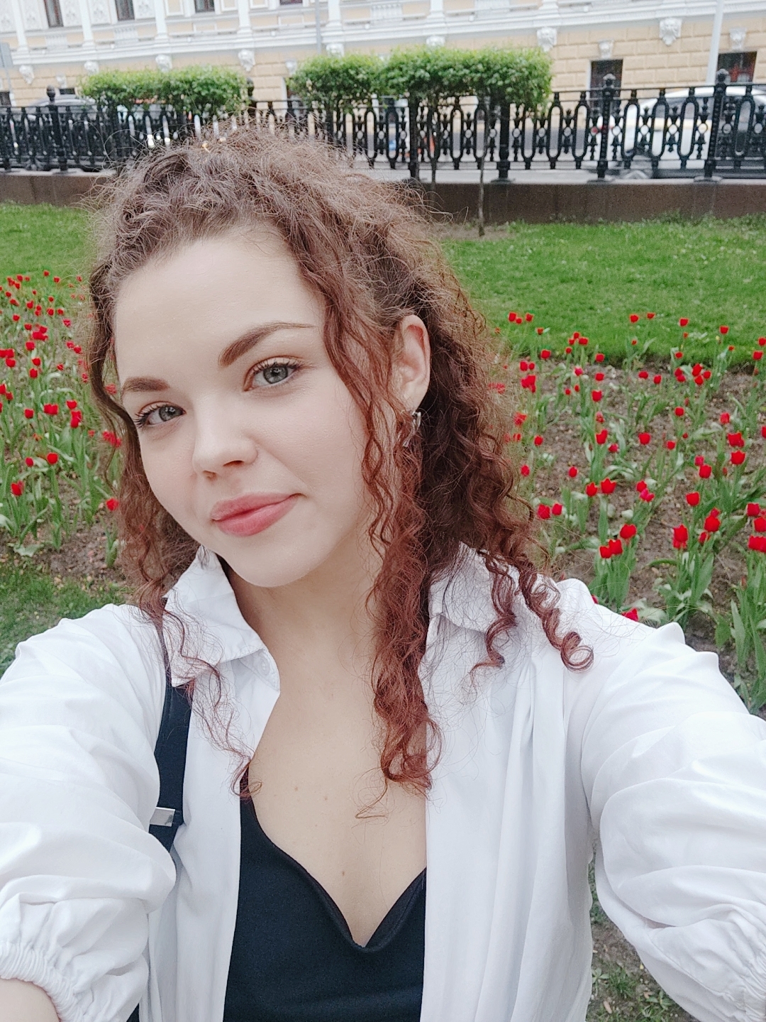 A woman with curly reddish-brown hair is taking a selfie outdoors in what appears to be a park setting. She is wearing a white, unbuttoned shirt over a black top. Behind her, a flowerbed filled with red tulips is visible. Further in the background, there's a green lawn and a black wrought-iron fence. Beyond the fence, a building with a light-colored facade can be seen. The presence of a Russian city building (with a typical facade) suggests this photo was taken in Moscow, Russia.