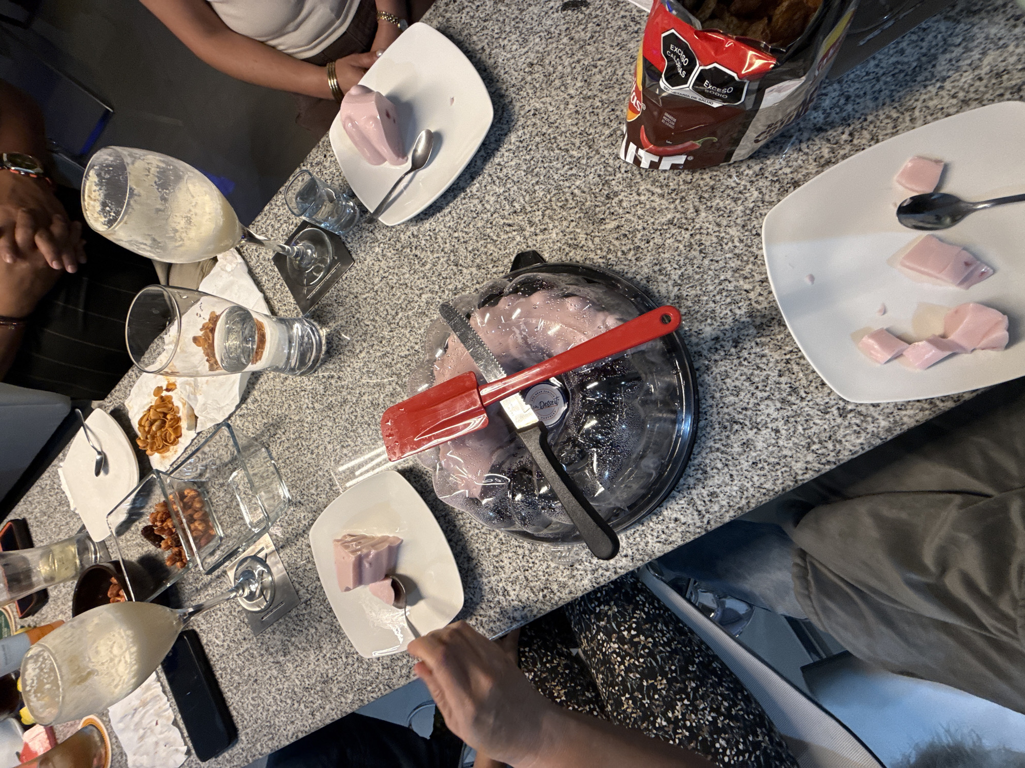 The image shows a tabletop scene with various objects related to food and drink, suggesting a gathering or meal taking place. The surface appears to be a gray speckled countertop. Scattered across the surface are plates containing what appears to be pink dessert items, potentially gelatin or pudding.
Several glasses, some partially filled with liquid, are present. There is a bag of chips labeled with the warning "Exceso Calorias Exceso Sodio," indicating high calorie and sodium content. Utensils, including spoons and serving implements, are laid out alongside the food. A covered dessert container with a red spatula and a knife rests on the table.
Partial views of people are visible, including hands and arms. The attire suggests casual wear. The environment suggests an indoor setting, with other seated individuals implied around the table. The location is not discernible from the image. Therefore, I cannot verify if this image was taken in Santa Clara Ocoyucan, Mexico.