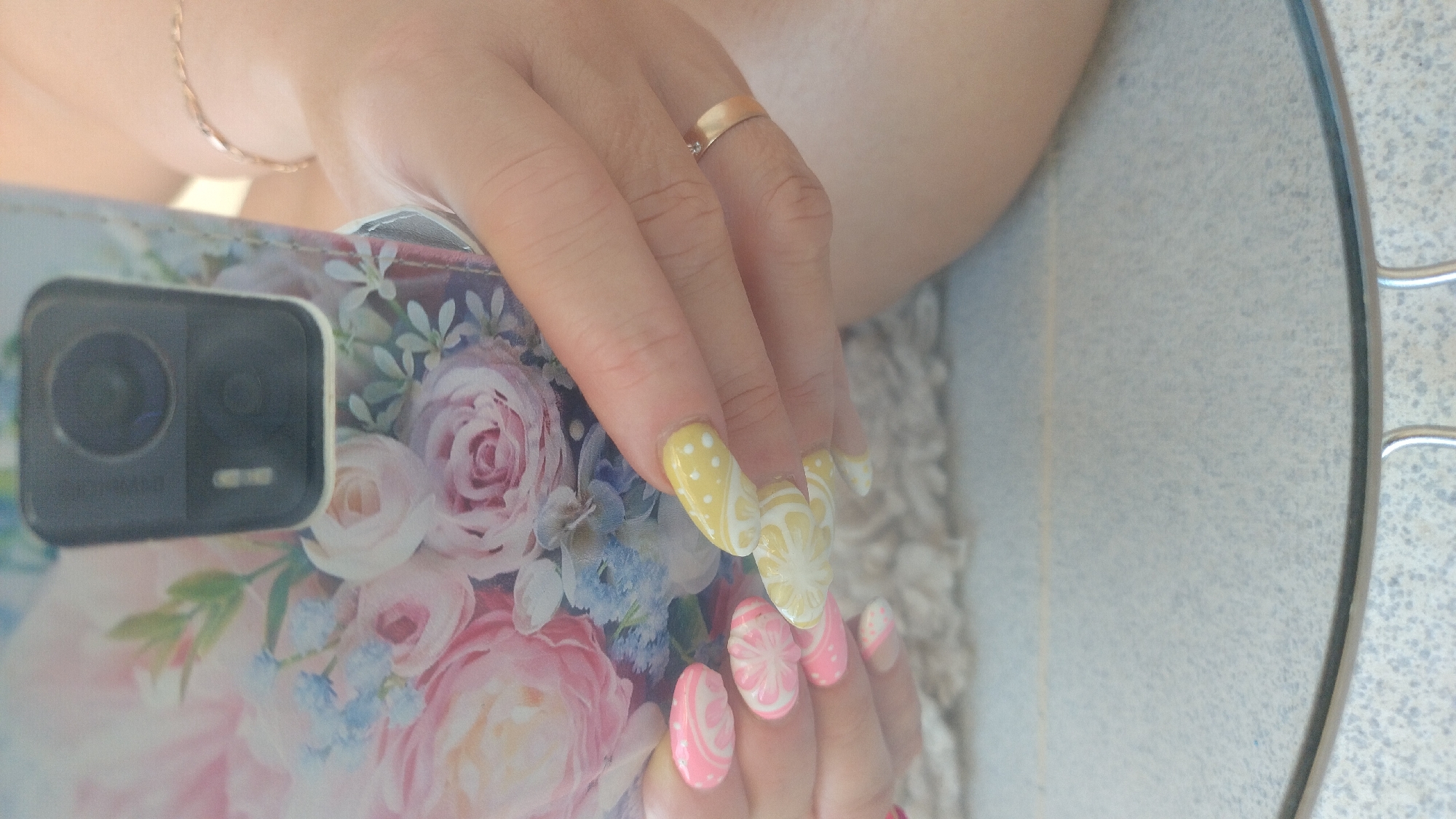 A close-up shot shows a person's hands holding a floral-patterned phone. The nails are painted with intricate designs, some in pink and white swirls and others in yellow with white polka dots and white citrus fruit patterns. A thin gold ring with a small stone is worn on the ring finger of the right hand. The background includes a blurred floral phone case, a fuzzy surface, and a tiled floor. The scene is indoors and appears to be a personal, relaxed setting. The location is not specified to be in Perea, Greece.