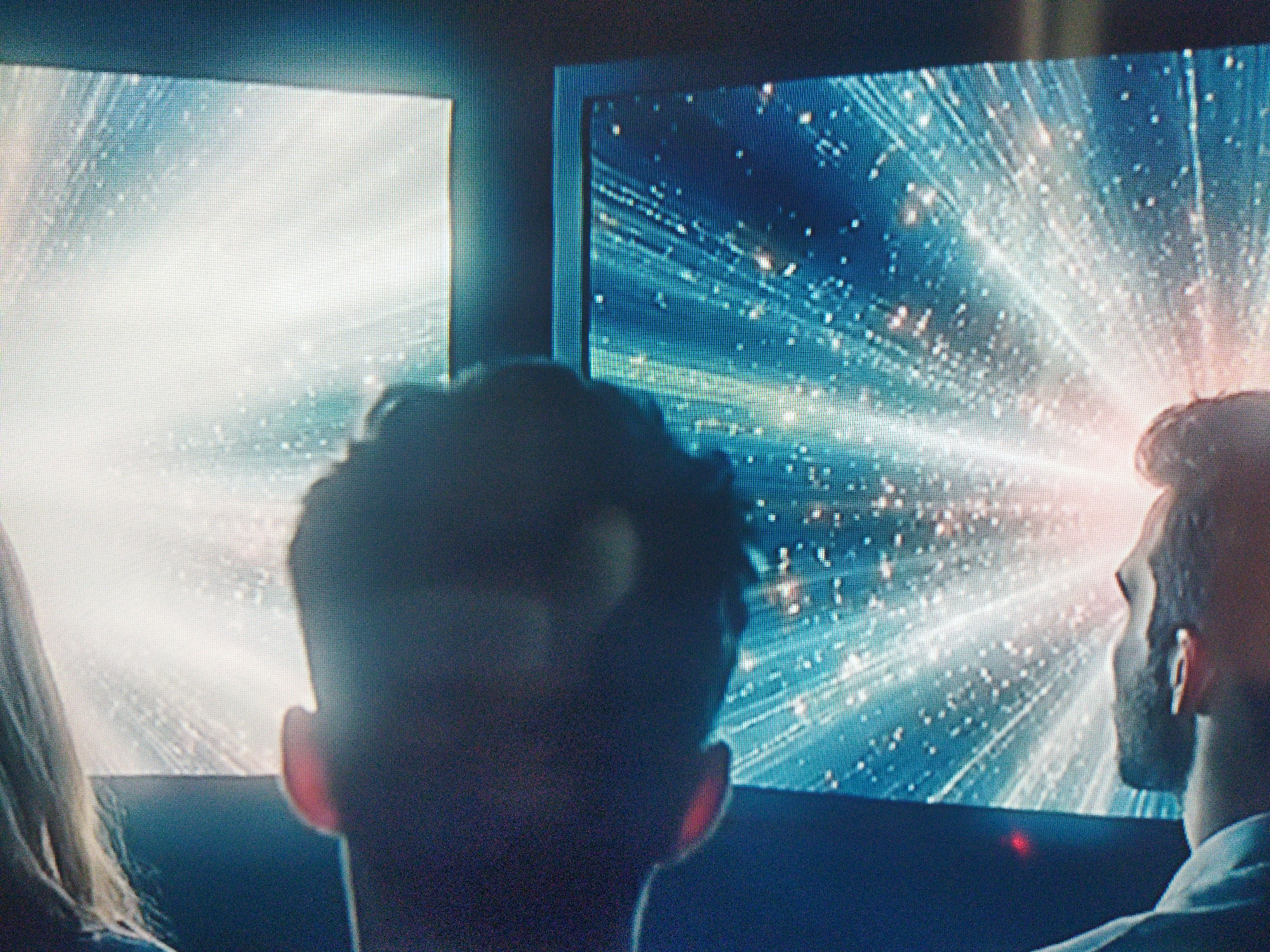 Two people are looking at two screens showing light effects. The screen on the left is white, while the one on the right has a blue background with white and orange rays. The location is not discernible in the image.
