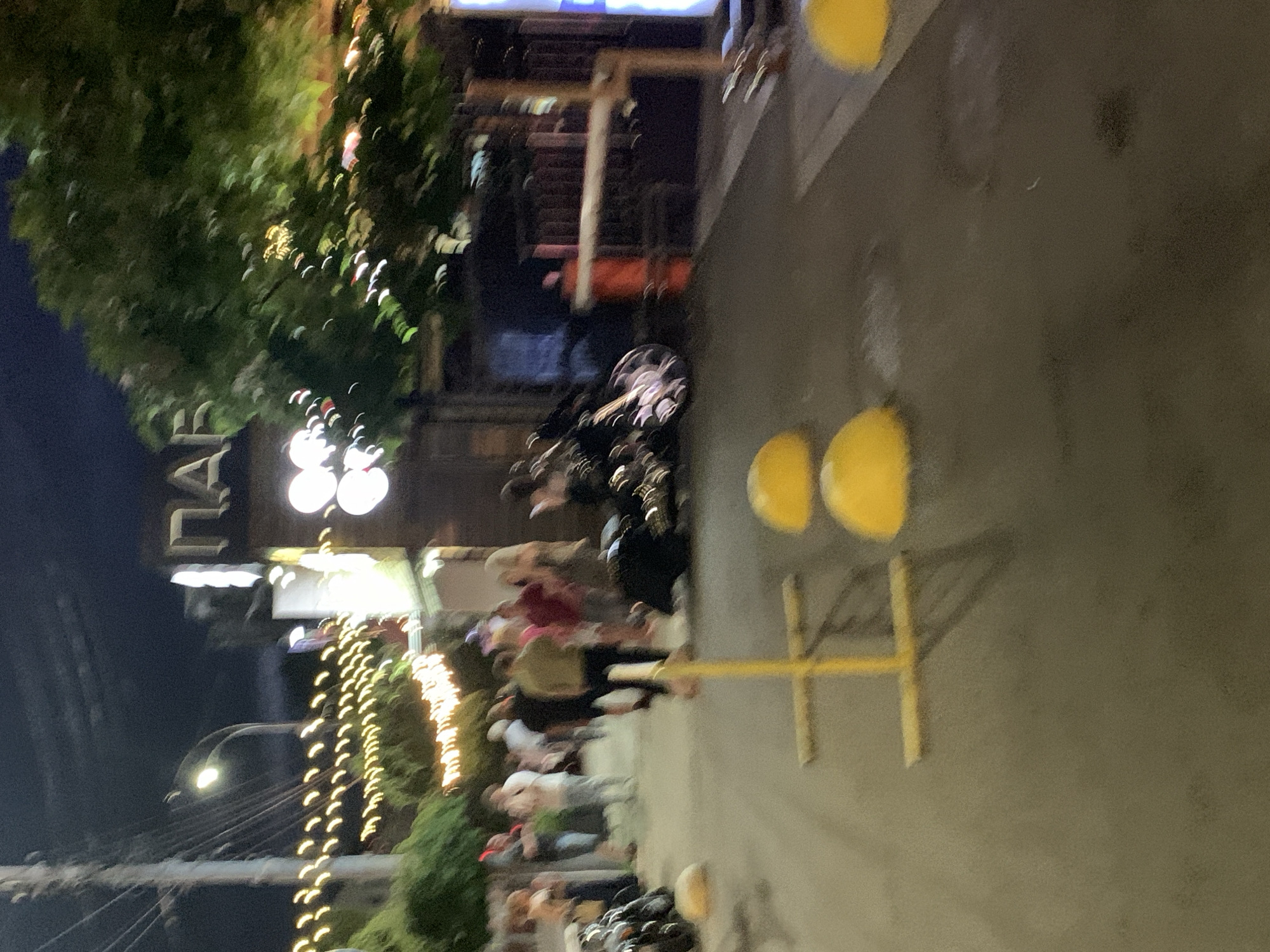 In the city of Unknown, Unknown, an outdoor scene is depicted at night. The foreground features an asphalt surface with yellow traffic delineators and two yellow speed bumps. Beyond this, a group of individuals are standing and walking, with several motorcycles parked to the left. In the background, a building with illuminated signage and a large tree are visible. String lights are draped across the scene, casting a warm glow. A yellow barrier arm extends across what appears to be an entrance or exit. The overall atmosphere suggests a lively, possibly commercial, area at night.