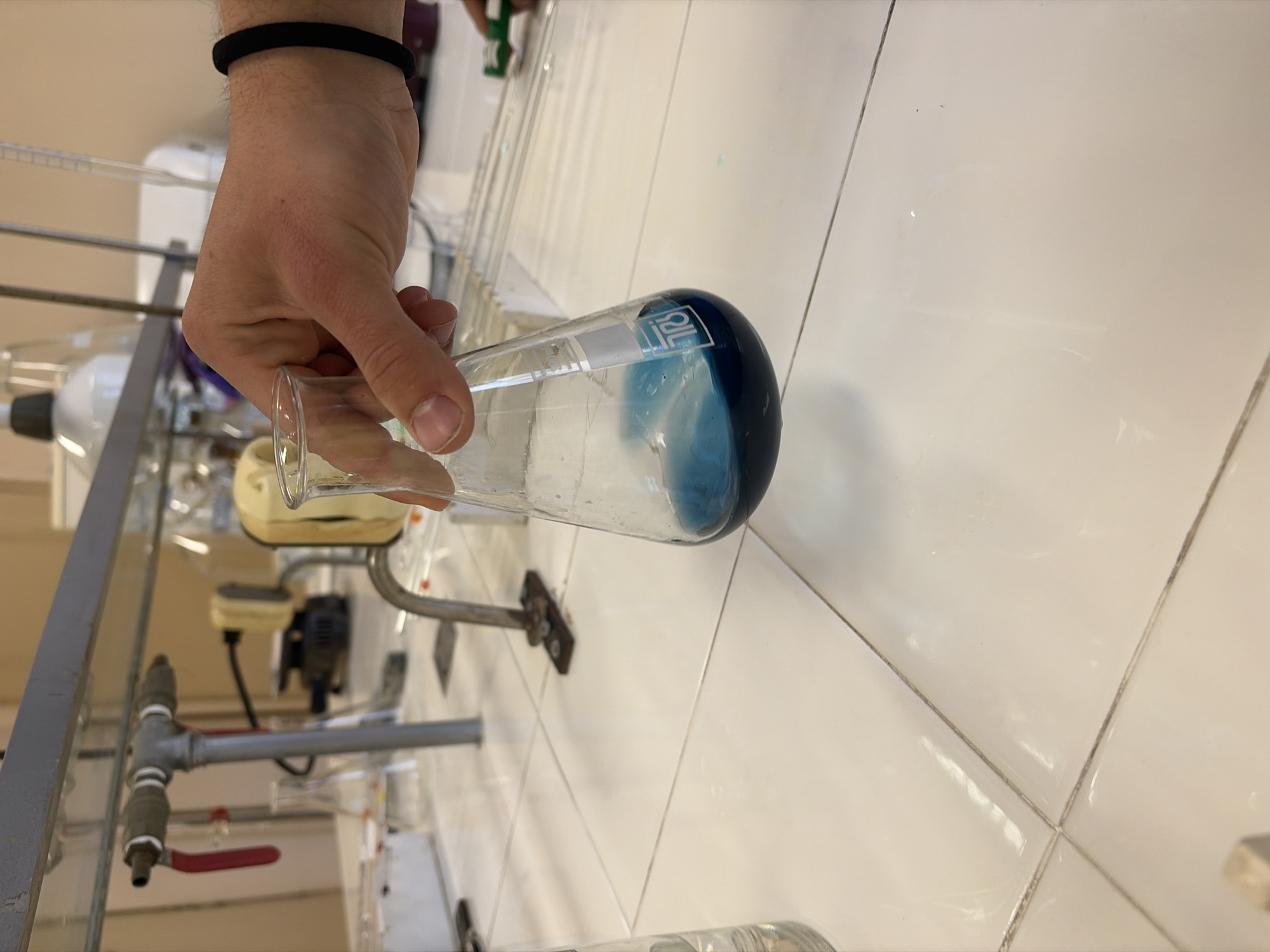 A person is holding a laboratory flask containing a blue liquid in a laboratory setting. The laboratory has white tiled walls and a gray metal frame supports various pieces of laboratory equipment, including a motor, tubing, and glassware. A red valve is visible on the equipment. The exact location and city are unknown.