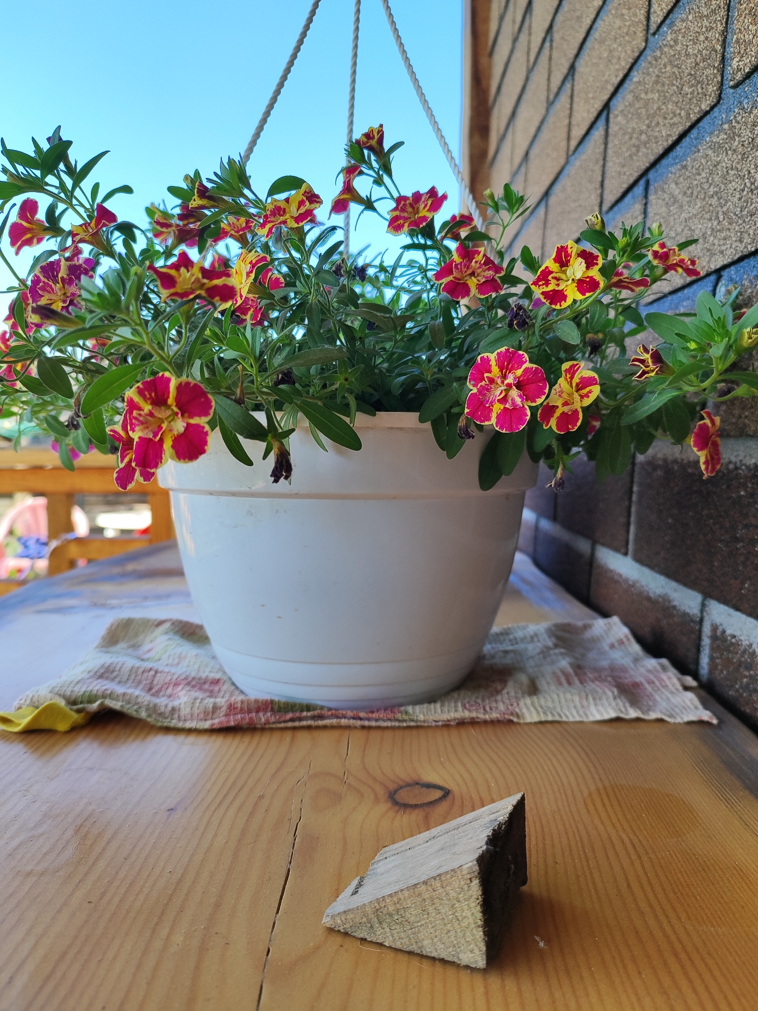 A close-up, eye-level shot captures a white hanging planter filled with vibrant pink and yellow petunias, suspended against a bright blue sky. The planter sits on a wooden table, partially covered by a patterned cloth. In the foreground, a small, angled piece of wood rests on the table's surface. The background features a textured, brown brick wall. The scene is set outdoors, likely on a porch or patio. No people are visible in the image.