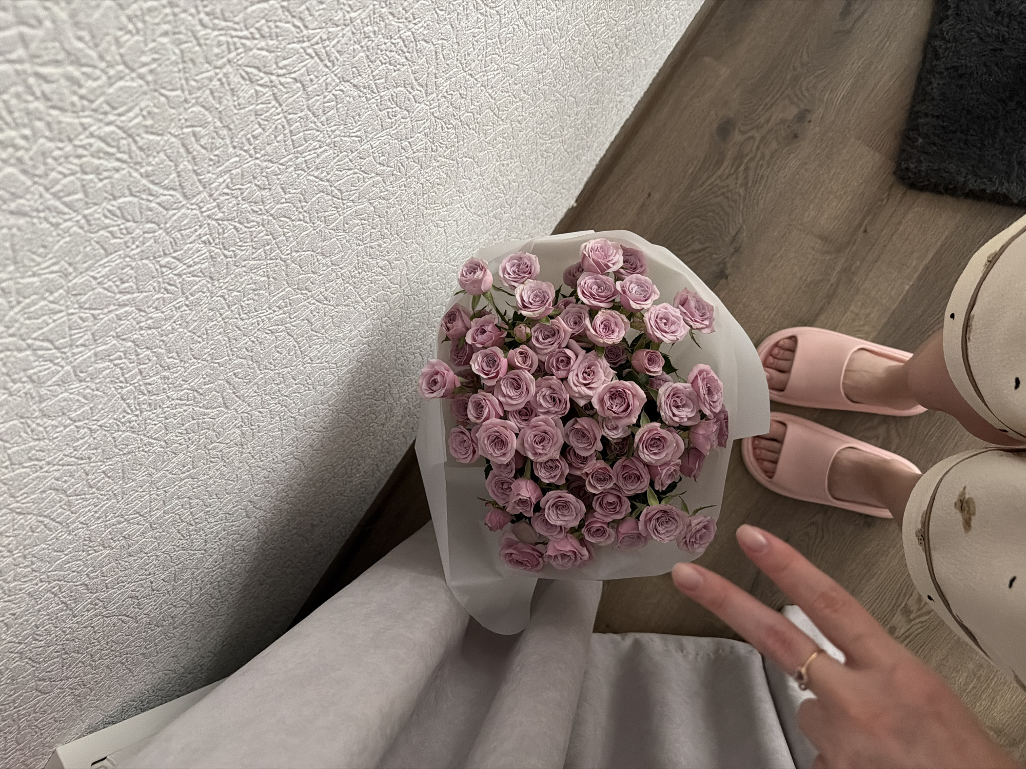An overhead shot shows a bouquet of pink roses wrapped in white paper, resting on a wooden floor. To the left of the bouquet, a hand displays a peace sign, adorned with a gold ring. Pink slippers are visible on a person's feet in the lower half of the frame. The background consists of textured white wallpaper and a light gray curtain. The scene is indoors, likely in a residential setting, with no discernible indicators of the city of Kyiv, Ukraine.