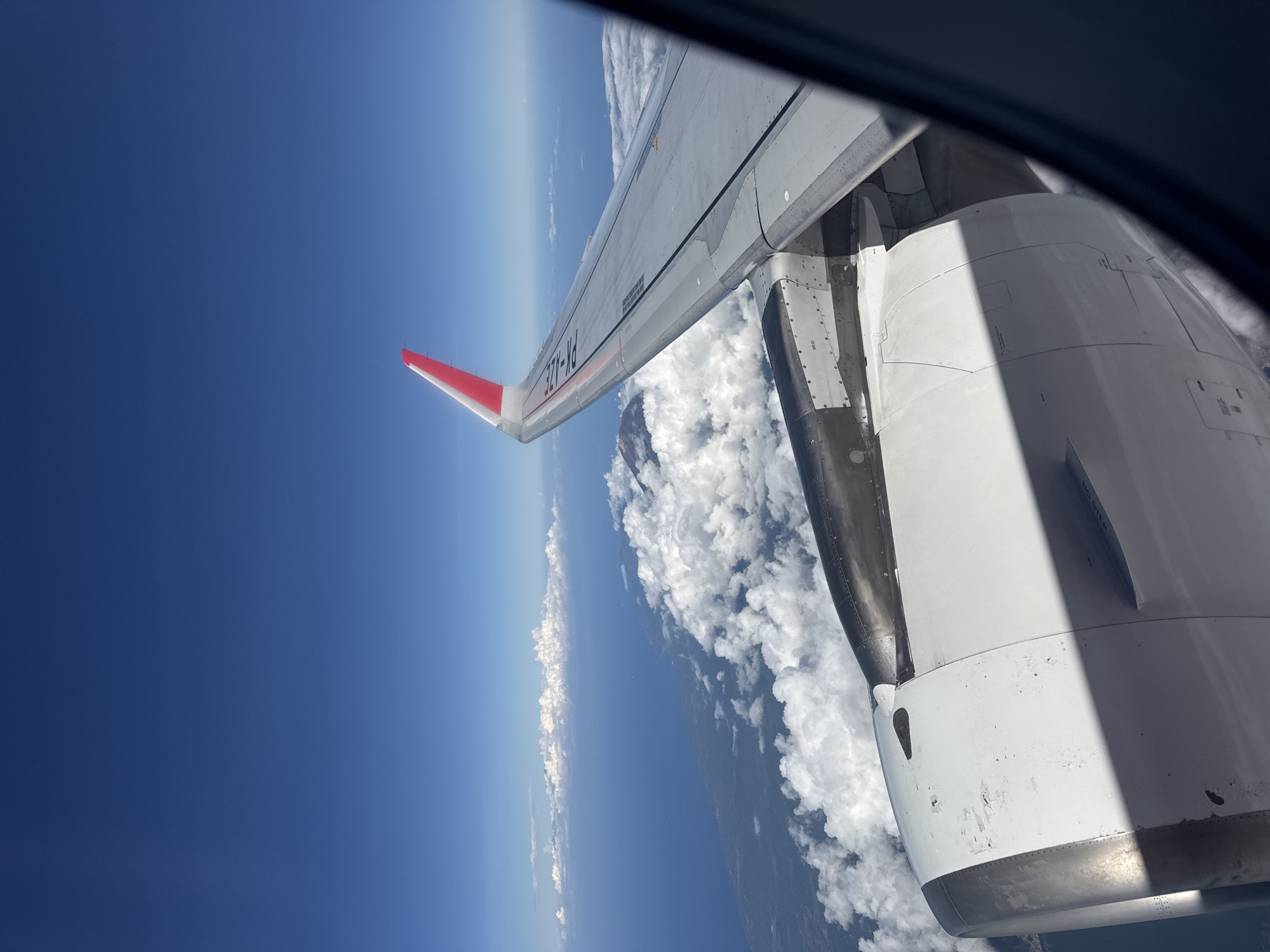 The image shows a view from an airplane window. The wing of the plane is visible, with a red winglet, and part of the engine is also visible. Below, there are clouds and a horizon line separating the sky from what appears to be the earth's surface. The sky is a deep blue. There is no indication the location is Bangli, Indonesia.