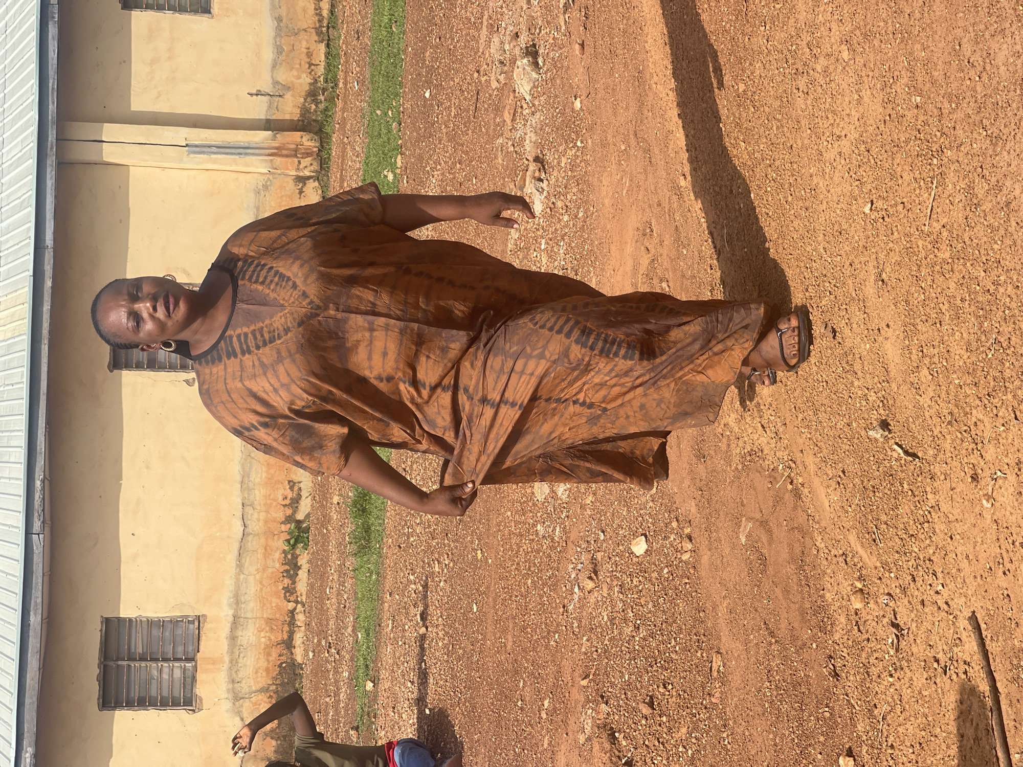 A woman stands on a dirt path next to a light-yellow building, likely in Akwanga, Nigeria. She is wearing a long, brown and black tie-dye dress and sandals. The building has a grey roof and barred windows. The ground is uneven and covered in small stones. The lighting suggests it is daytime.