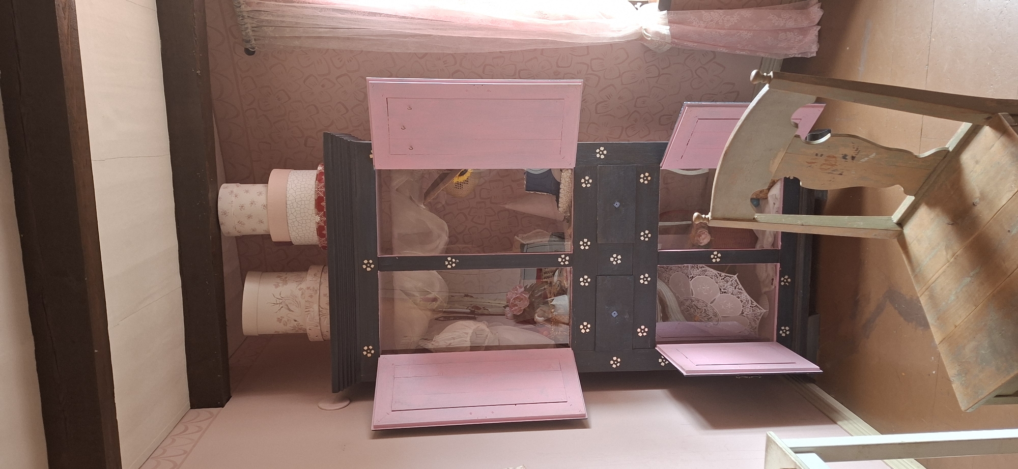 A wooden chair with a light blue-green painted finish sits in the foreground of a room. The chair has a rectangular seat with wooden planks and a decorative, curved backrest. Behind the chair is a dark gray cabinet with pink doors. The cabinet has multiple compartments and drawers, some of which are open, revealing decorative items like lace and fabric. On top of the cabinet are stacked hat boxes adorned with floral patterns. The room is painted in soft pink tones, and a sheer pink curtain hangs on the right side of the image. The floor is a reddish-brown color. The overall scene suggests a vintage or cottage-core aesthetic. The location is not specified in the image or its context.