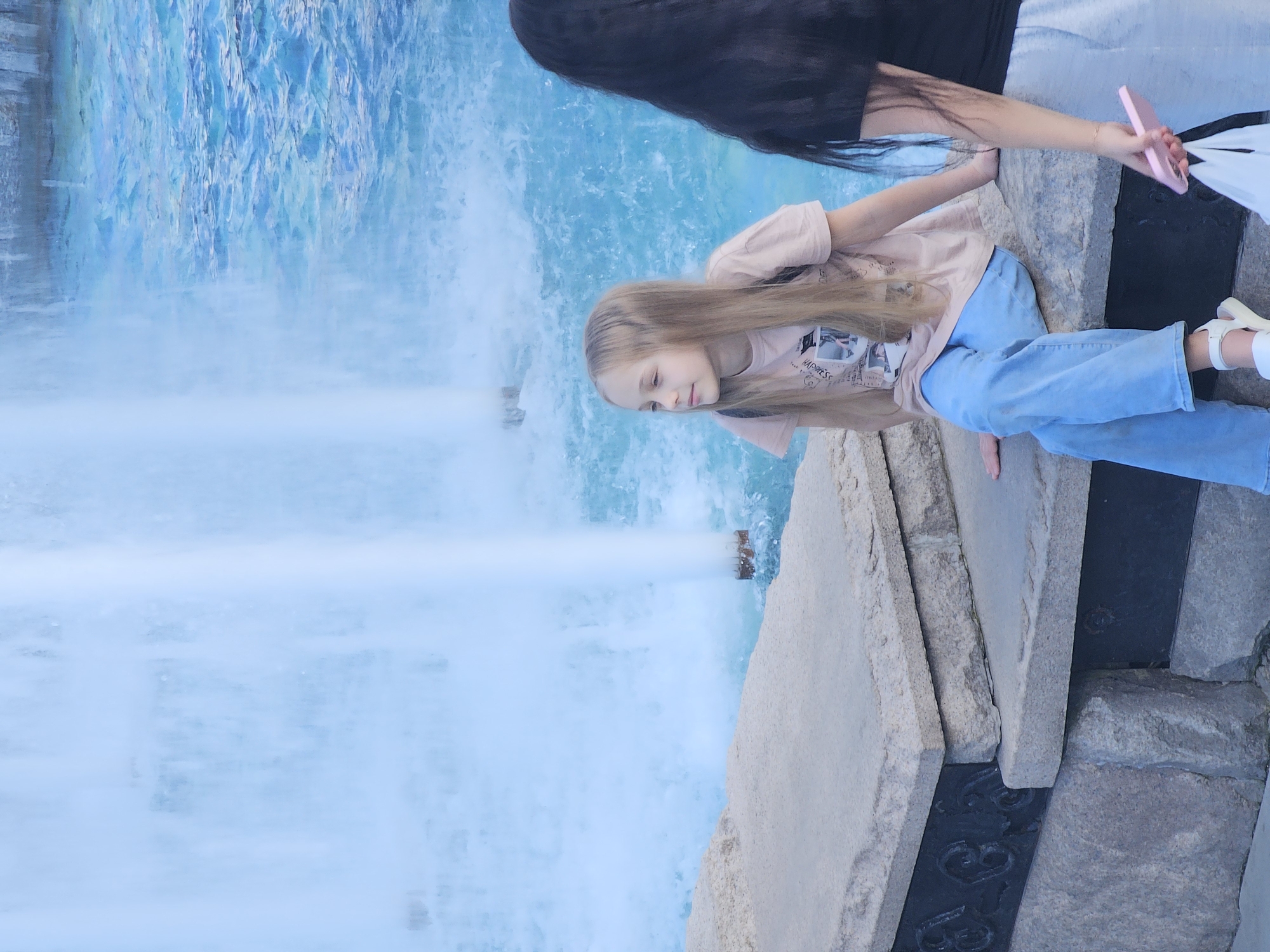 The image, if rotated 90 degrees clockwise, displays a young girl and a partially visible adult alongside a large, active fountain or artificial waterfall during what appears to be daytime.
The young girl, with long, light brown hair, is positioned in the foreground. She is wearing a light-colored (possibly beige or light pink) t-shirt and light blue jeans, paired with white sandals. She is leaning forward, peering intently into the cascading water, her expression calm and focused.
Behind the girl and to her right, an adult is partially visible. Only their long dark hair, the shoulder of a dark top, and one hand holding a pink smartphone are clearly seen.
The water feature is prominent, showcasing vibrant blue water at the top that dramatically falls as a wide, white, and frothy cascade. The falling water creates a mist and lighter blue, foamy water at its base. The people are standing on or next to a substantial structure made of large, light-colored stone blocks. Between some of these blocks, particularly towards the bottom right, dark, possibly metal elements with decorative, ornate patterns resembling the letter 'B' or number '8' are visible.
On the girl's t-shirt, faint text can be seen, which includes the word "HAPPINESS". The overall scene suggests a leisurely moment of observation at a public water display.