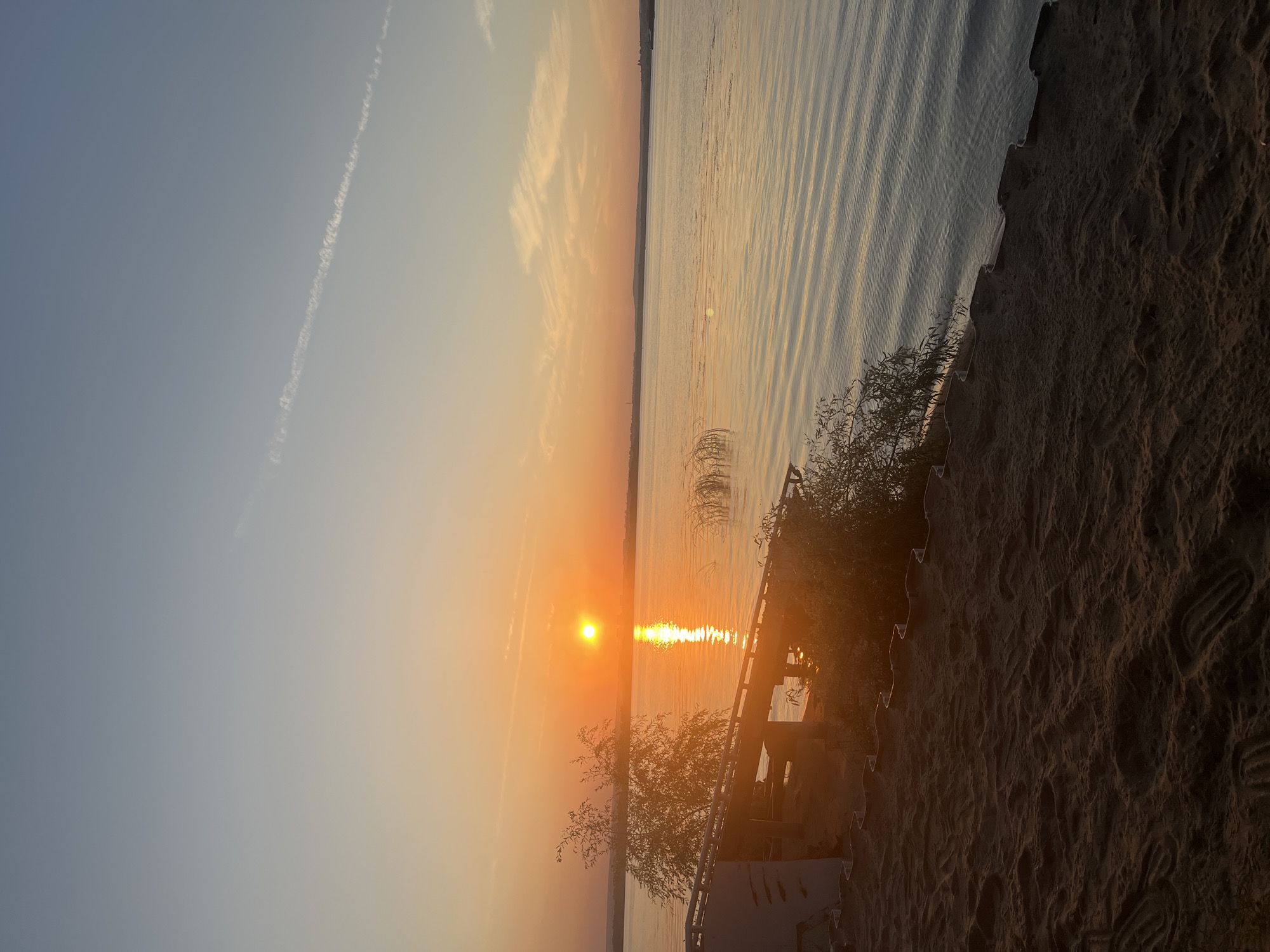 An outdoor vertical shot captures a sunset over a body of water, likely the Volga River, near Shumeika, Russia. The sun, a bright orange orb, is low in the sky, casting a golden reflection on the rippling water. The sky transitions from a pale blue at the top to a warm orange near the horizon. A thin contrail stretches across the upper sky. In the foreground, a sandy embankment slopes down to the water's edge. On the embankment, a wooden structure with an angled roof is visible, partially obscured by greenery. Some reeds grow near the water. The sand shows numerous footprints. The scene is peaceful and serene, depicting the end of a day.