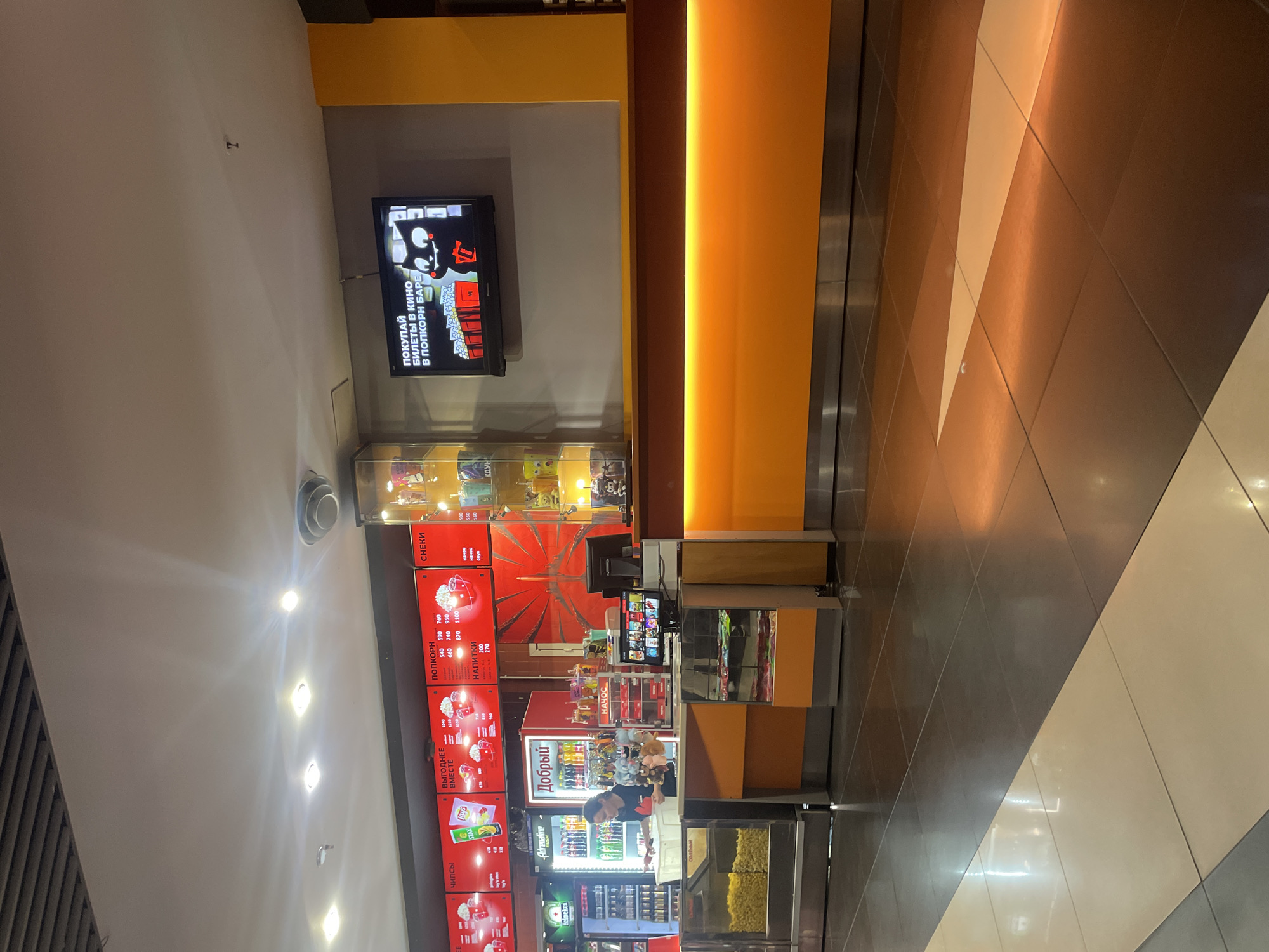 The image depicts a concession stand in a cinema lobby. The stand features a bright orange counter with a glowing light strip underneath. Above the counter, red menu boards display prices for various items like chips, popcorn, and drinks. A refrigerator unit stocked with beverages is visible to the left, with a young woman in a dark shirt working behind the counter. She appears to be attending to a customer or preparing an order. To the right, a flat-screen television mounted on the wall shows an advertisement featuring a cartoon black cat and the text "ПОКУПАЙ БИЛЕТЫ В КИНО В ПОПКОРН БАРЕ," which translates to "BUY MOVIE TICKETS AT THE POPCORN BAR." Display cases near the counter contain packaged snacks and merchandise, including plush toys. The floor is tiled with a combination of dark grey and lighter beige tiles, reflecting the ambient lighting. The overall environment suggests a commercial space designed for leisure, likely a cinema in Vaganovo, Russia.