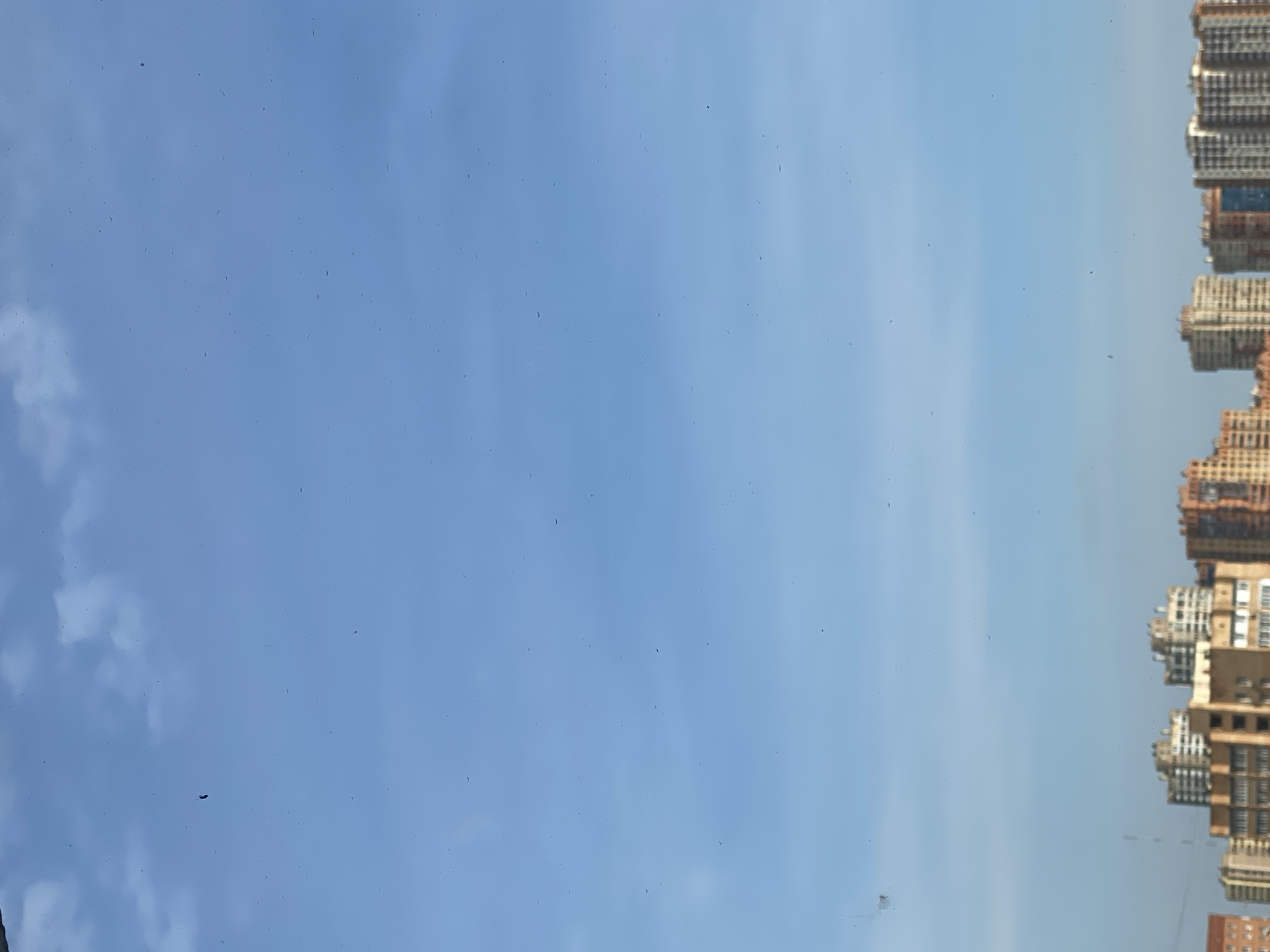 This is a vertical, medium shot of a clear blue sky with some wispy clouds. In the lower portion of the image, the tops of several apartment buildings are visible, creating a city skyline. The buildings are of varying heights and architectural styles, with some appearing to be modern high-rises and others more traditional apartment blocks. The sky is bright and sunny, suggesting a daytime scene. There are some small specks visible on the lens, which appear as dark dots scattered across the image. One of these specks is a single black comma-like shape in the upper left quadrant of the sky.