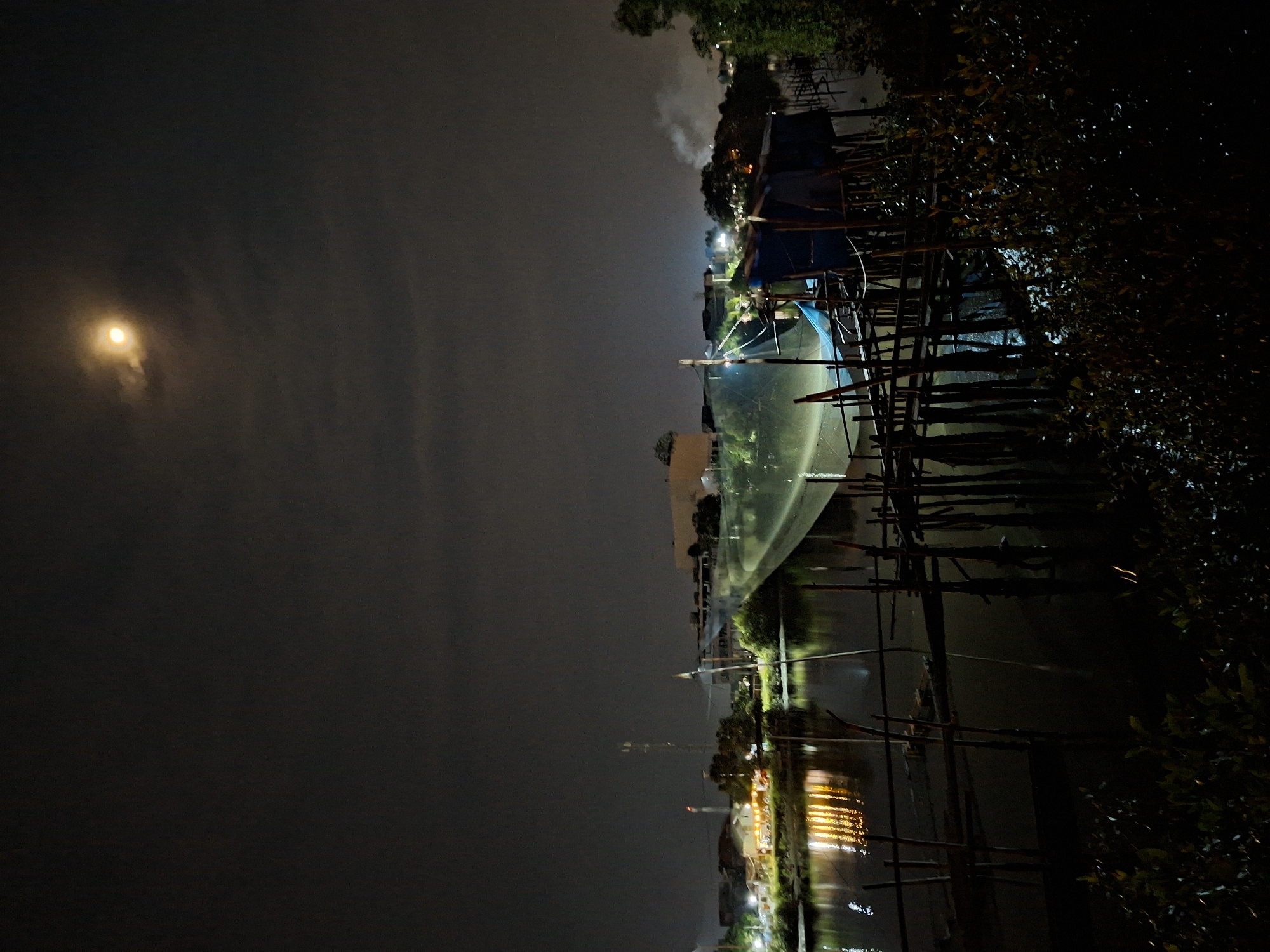 At night in Lhokseumawe, Indonesia, a waterway reflects lights from buildings and a telecommunications tower across the water. A crescent moon is visible through the overcast sky. A fishing boat is docked on the waterway, partially covered with a blue tarp. Support structures made of dark poles are installed along the dock and obscure some of the view, and some branches of trees and other foliage are visible in the far right. The scene is dark with reflections off the water.