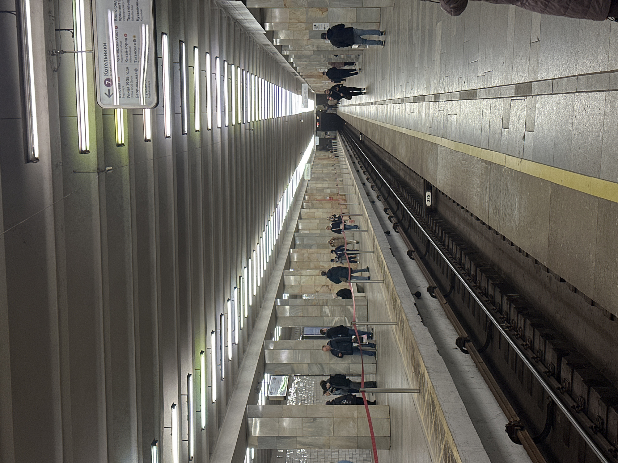The image depicts a subway station, presumably in Moscow, Russia. The location appears to be the Kotelniki station on Line 7, based on the sign visible on the left. The station features a long platform with fluorescent lights along the ceiling. Several people are on the platform, some walking and others standing. A track runs along the edge of the platform. The platform surface has tiled floors, and there are posts with red ropes sectioning off the lower levels.