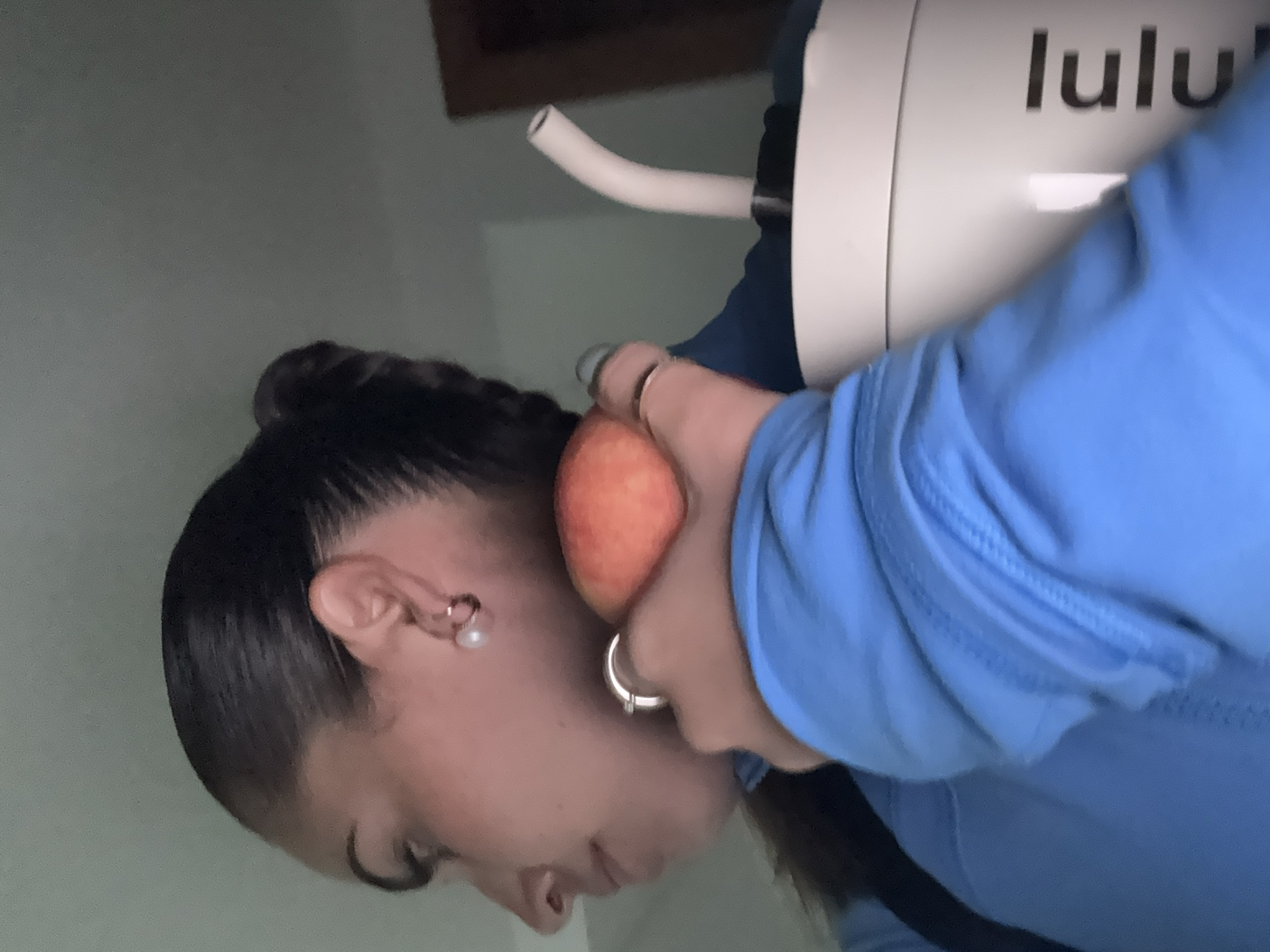 A woman with dark hair pulled back in a braid is seen in a side-view, holding an orange peach close to her face with her right hand. She is wearing a blue shirt. She has a silver hoop earring in her ear. Next to her is a white water filtration device. The word "lulu" is visible on the device. The location of the image is not discernible from the image.