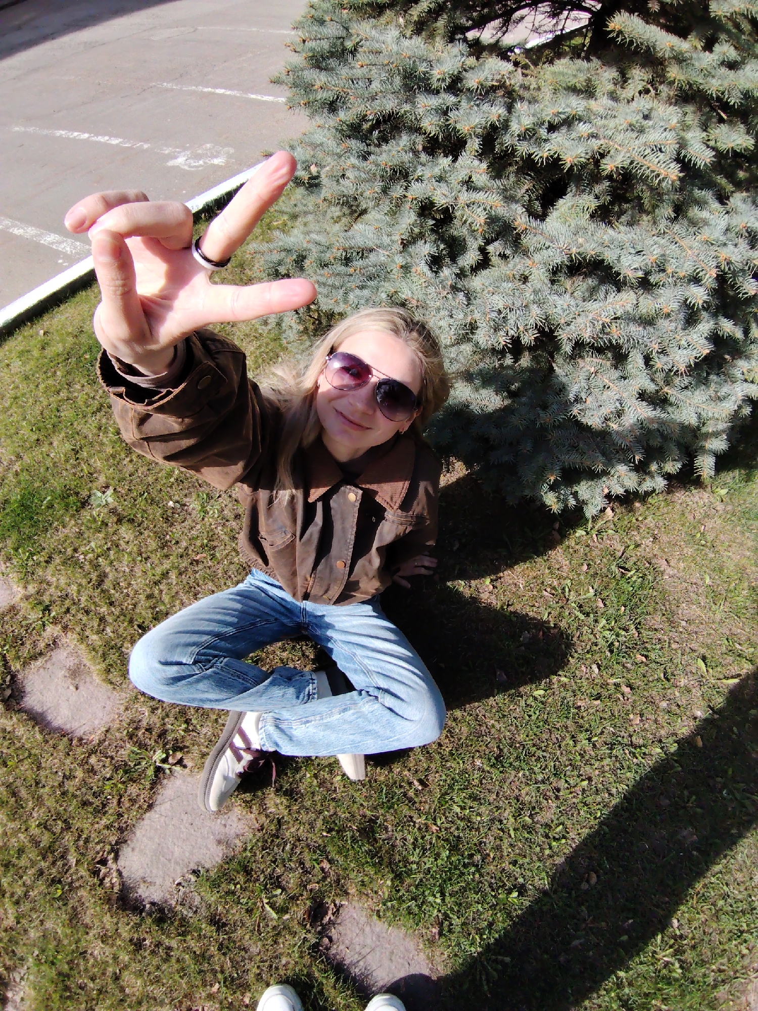 A person sits cross-legged on grass near a blue spruce tree. The person wears a brown jacket, blue jeans, sunglasses, and sneakers, and is displaying a hand gesture with two fingers extended, rings on the fingers. The location appears to be an urban area, specifically the city of Chaikovsky, Russia, as deduced from prior knowledge and the visual context suggesting an outdoor setting with a paved area in the background.