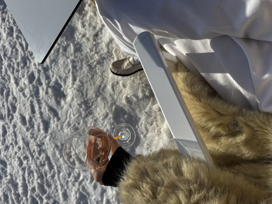 An overhead, first-person perspective captures a person seated outdoors in Courchevel, France, on a sunny day. The scene is dominated by a field of textured white snow under bright illumination. The subject's right hand, adorned with light-colored fingernail polish, holds a clear stemmed wine glass containing a rosé-colored liquid and a partially visible orange stirrer. The individual wears a light brown or tan fur-like coat, visible on the left side of the frame with a metal snap closure, and white or light-colored trousers on the right. A single light beige loafer is visible on the right foot. A white rectangular armrest of a chair spans horizontally across the lower middle of the image. A dark, possibly black, tabletop is partially visible in the upper right, suggesting an outdoor dining or lounge area.