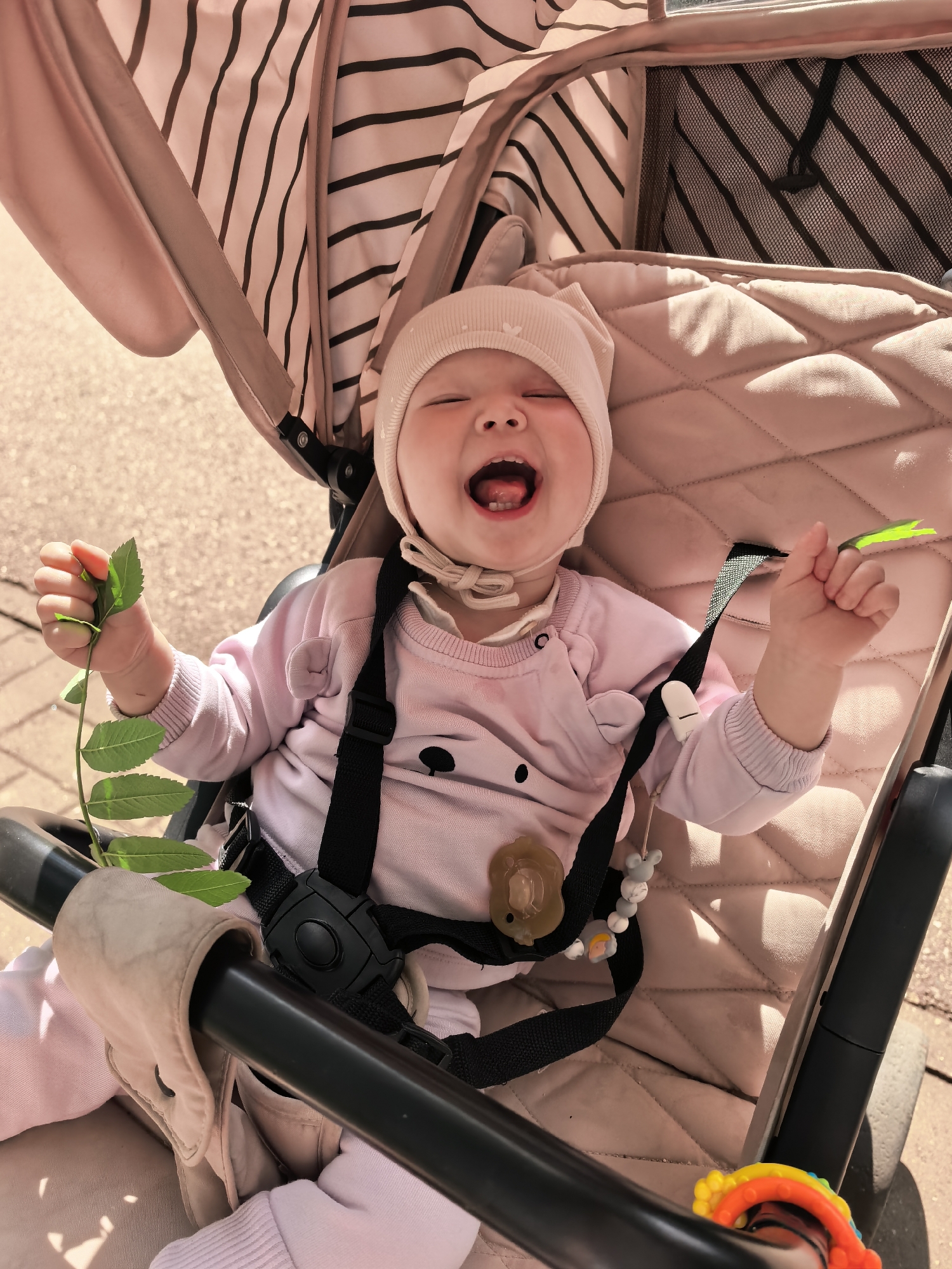 A close-up shot shows a baby sitting in a stroller, laughing. The baby, wearing a pink onesie and a matching bonnet, is holding a small green plant in their left hand and a bright green object in their right. A pacifier is clipped to the stroller's harness, along with a beaded teething toy. The stroller itself is a neutral beige color with a striped canopy. The background appears to be an outdoor setting, with paved ground visible, suggesting a park or a street. The lighting indicates it is daytime. The image does not contain any elements that would allow for identification of the location as Sterlitamak, Russia.
