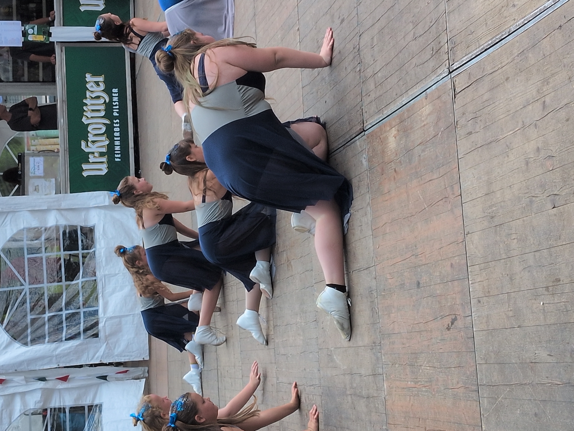 In Rastenberg, Germany, a group of young women in matching dance attire are performing on a wooden stage. The stage is set up outdoors, with a white tent structure visible in the background. The women are dressed in navy blue skirts and grey leotards, with blue hair accessories. They are in various kneeling and crawling positions, suggesting a choreographed dance routine. The scene appears to be a public event or performance, possibly a festival or competition. The background shows a green banner with white lettering that reads "Urkrostitzer Fein Herbes Pilsner," indicating a sponsorship or branding associated with the event. The overall impression is one of synchronized movement and focused performance in a festive outdoor setting.