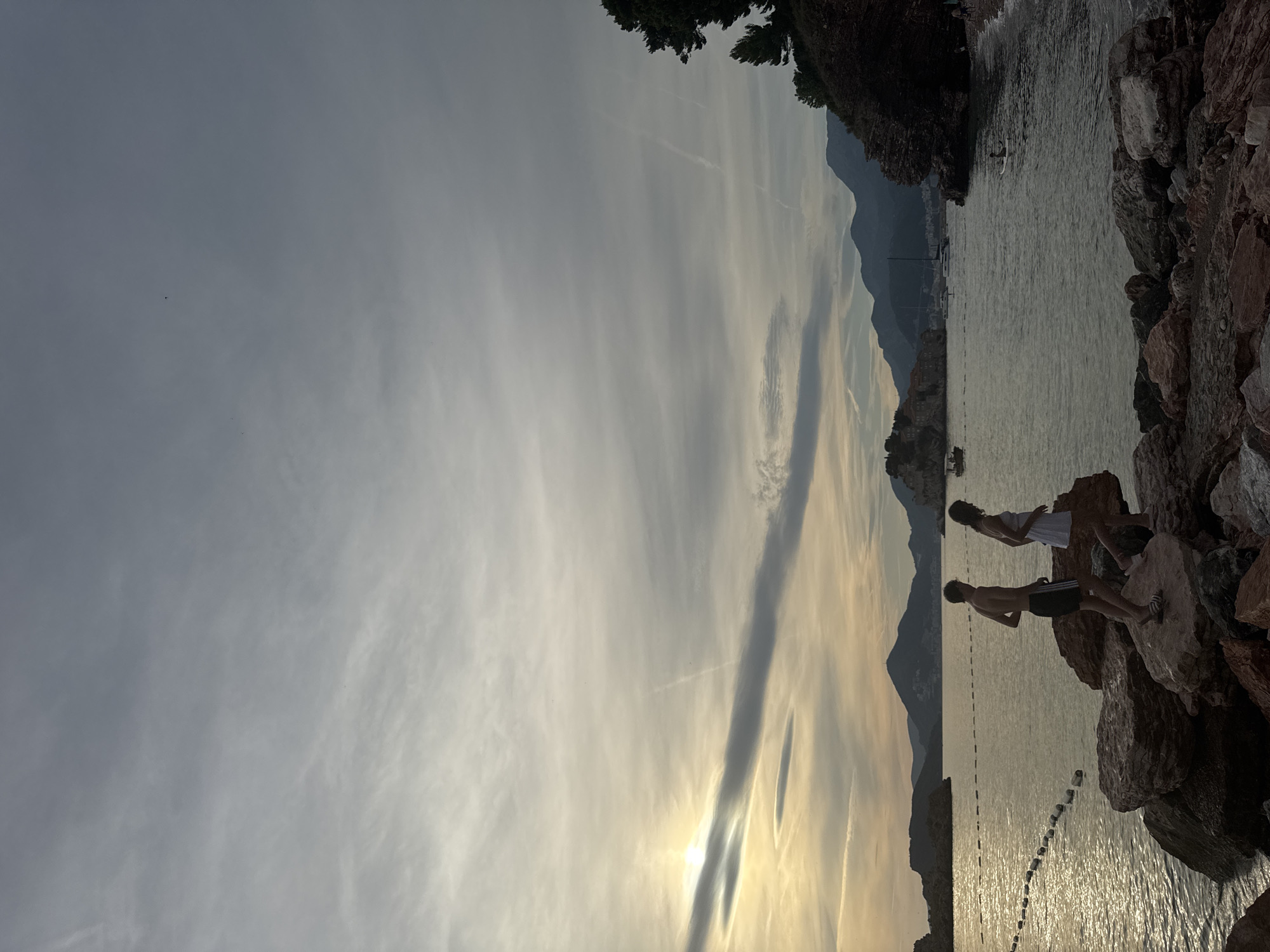 In Kamenovo, Montenegro, two individuals, a male and a female, walk across a rocky shore overlooking a body of water. The male, wearing black shorts, is positioned to the left, while the female, in a white dress, is to his right. They are positioned close to the water's edge. The sea is calm, with gentle waves lapping against the shore. In the distance, islands with buildings and lush vegetation are visible, along with sailboats and a small boat on the water. Mountains form a backdrop to the scene, their peaks shrouded in a soft haze. The sky is overcast with diffused sunlight peeking through the clouds, casting a serene, ethereal glow over the landscape. The overall atmosphere is tranquil and peaceful.