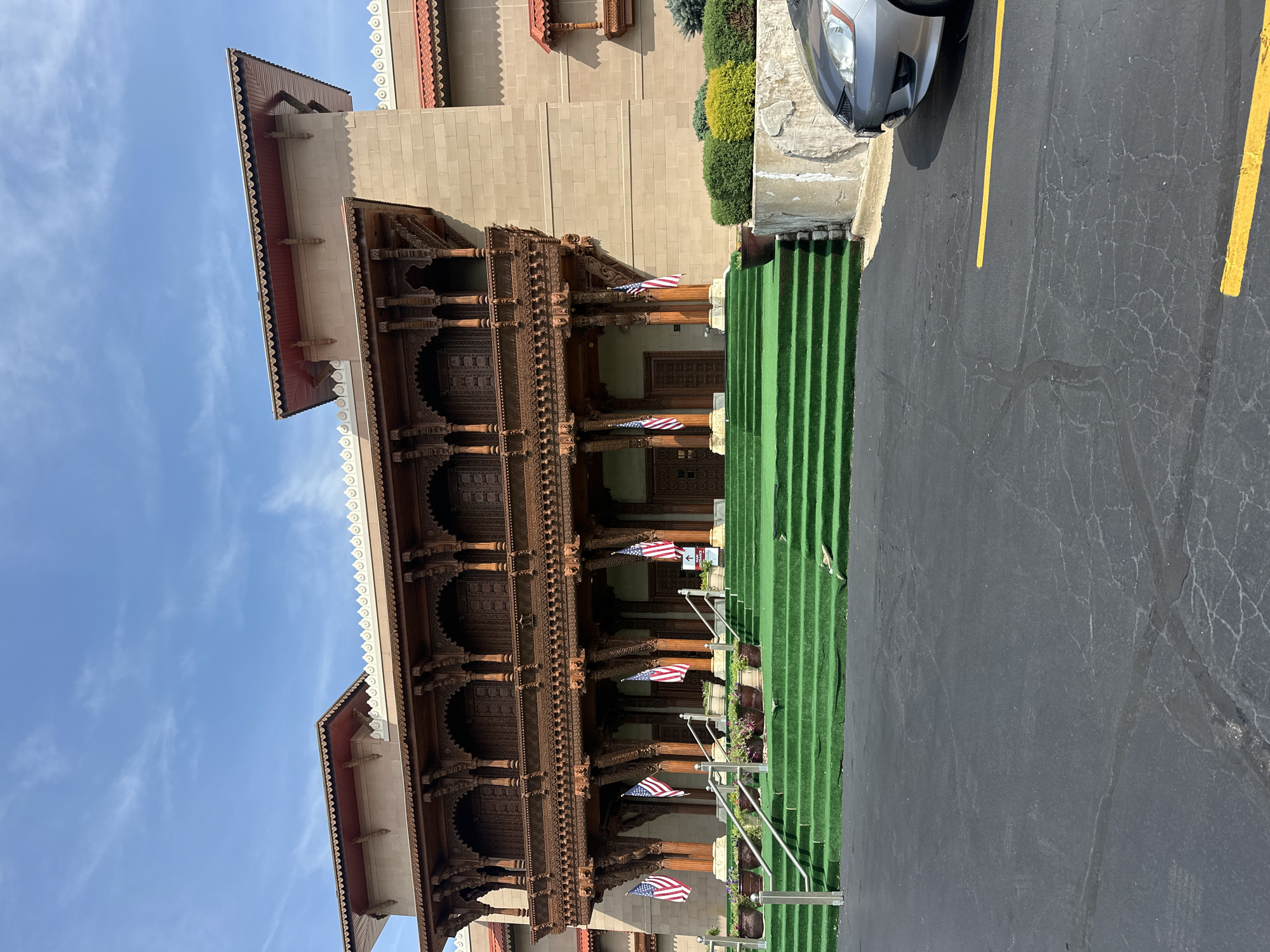 The image shows the exterior of a large, ornately decorated building with a tan stone facade and a prominent wooden balcony. The entrance features a wide set of stairs covered in green artificial turf, with metal handrails on either side. American flags are displayed along the staircase and at the entrance. The building has a distinct architectural style, with intricate carvings on the wooden elements. In the foreground, there is a black asphalt parking lot with yellow lane markings. A silver car is parked to the right of the frame. The sky is blue with scattered white clouds. The building is likely a temple or cultural center, given its elaborate design and the presence of flags. This is the BAPS Shri Swaminarayan Mandir located in Bartlett, Illinois, a suburb of Chicago in the United States of America.