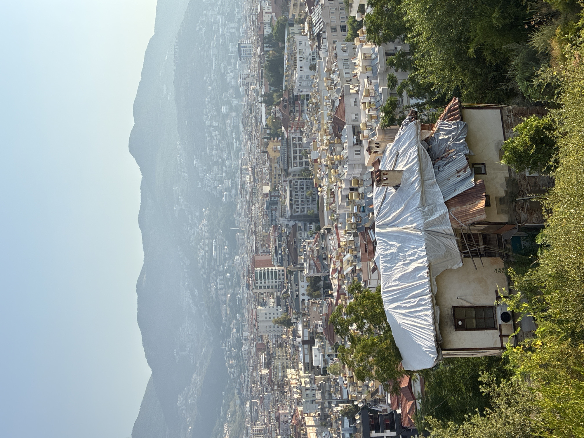The image shows an elevated view of the city of Alanya, Turkey, with a foreground focus on an old house covered with a tarp. The house has a damaged roof made of corrugated metal and tiles, and appears to be in a state of disrepair. An air conditioning unit is visible on the exterior wall. Behind the house, a densely populated urban landscape unfolds, characterized by numerous buildings with flat roofs, many of which are equipped with solar water heaters. The city stretches towards a hazy mountain range in the background. Lush green trees and vegetation surround the foreground house and also dot the urban landscape. The sky is clear and pale blue, indicating daytime.