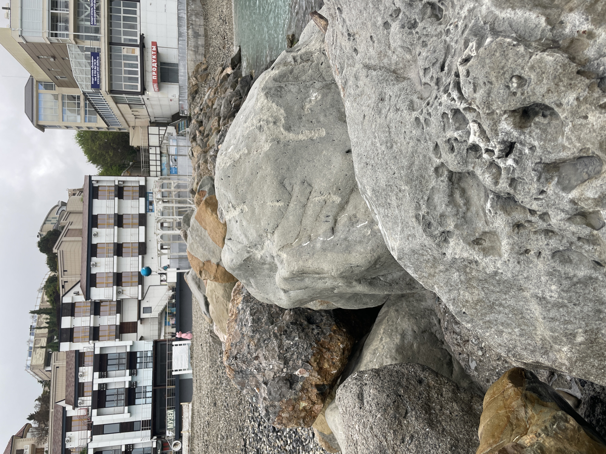 The scene is located in Olginka, Russia. Large, gray rocks dominate the foreground. A building complex stands in the background, featuring a modern, multi-story structure with black-and-white checkered designs. Other buildings are visible, with various architectural styles, including glass and concrete. A blue ball hangs near the checkered building, and a pink object sits on the ground. The sky is overcast.