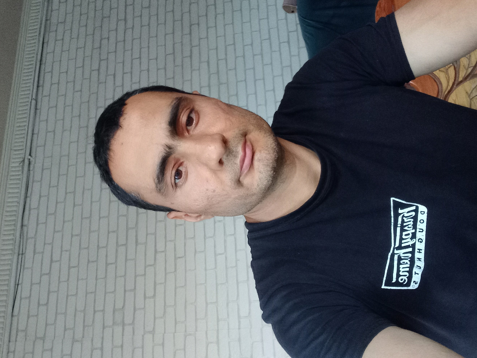 The media file is a portrait of a man, likely taken indoors. The man is looking directly at the camera with a neutral expression. He has short, dark hair and dark eyes. He is wearing a black t-shirt with a white logo on the left side of his chest. The logo appears to be a rectangular box with stylized text inside. The background consists of a light-colored brick wall pattern. The image is oriented vertically, suggesting it might be a selfie or a photo taken from a low angle.
The visible text on the t-shirt reads "JOUGHN DOUGHE 2", though the exact nature of the brand or message is unclear due to the text's orientation and possible stylization.
While the specific activity is not evident, the framing and pose suggest a casual, personal photograph. The lighting appears to be artificial, consistent with an indoor setting. There are no clear indicators of the time of day or weather. Given the provided location context of Samarkand City, Uzbekistan, the brick wall pattern might reflect local architectural or decorative styles.
