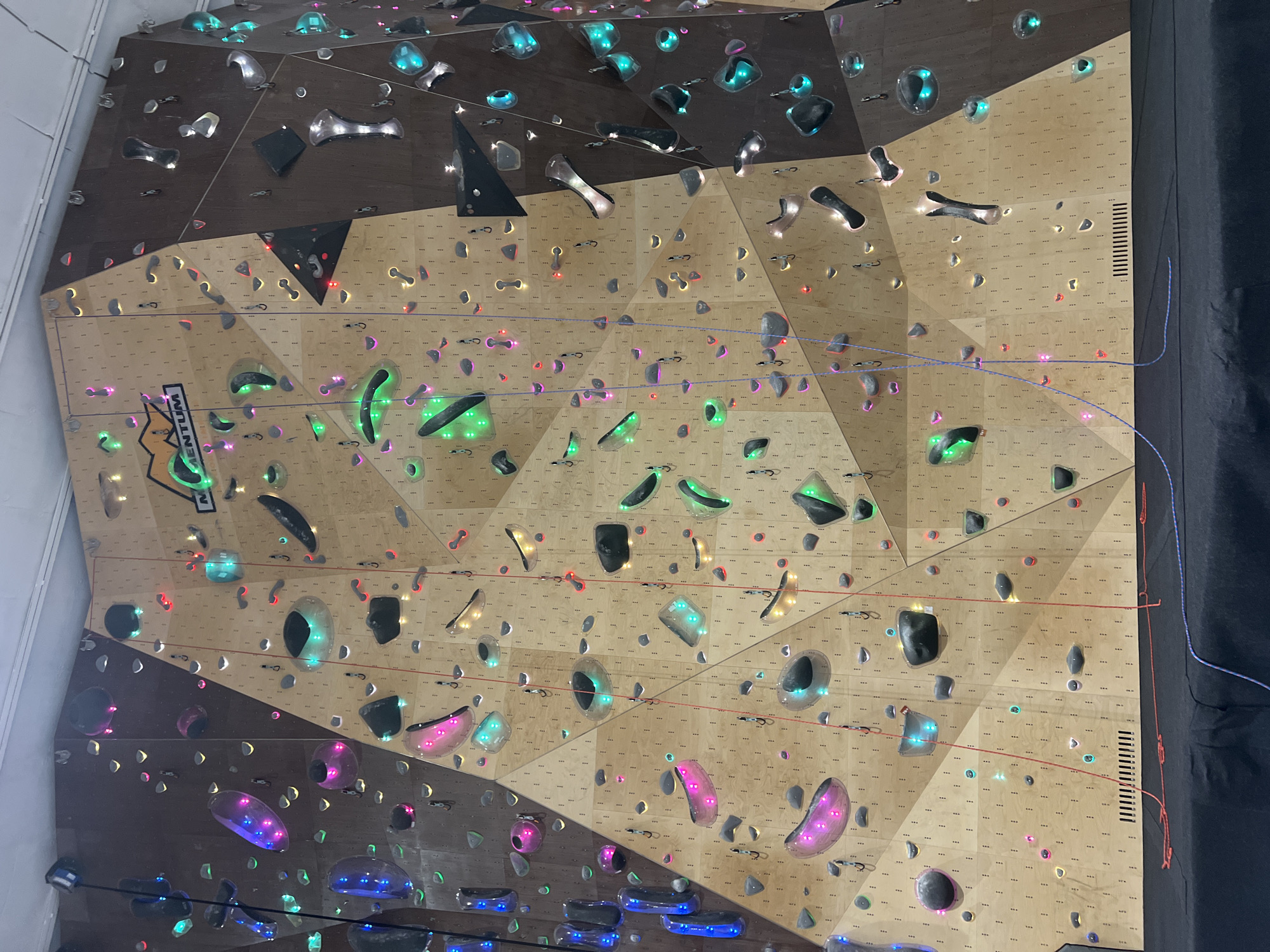 This image depicts an indoor climbing wall at Momentum climbing gym, located in Plovdiv, Bulgaria. The large wall is constructed from geometrically arranged panels of varying shades of wood, primarily light brown plywood and darker brown engineered wood. Numerous climbing holds are affixed to the wall; many are transparent or translucent and internally illuminated with LED lights displaying diverse colors such as pink, green, blue, red, and yellow. Other holds are opaque, appearing in black, grey, or metallic silver tones. Several climbing ropes, predominantly red and purple/blue, hang vertically down the wall from anchors at the top. The base of the climbing wall is covered by a thick, black padded mat designed for impact absorption. Horizontal slatted ventilation grilles are visible along the bottom edge of some light brown wall sections. The ceiling, partially visible at the top, is white. No individuals are present in the scene.