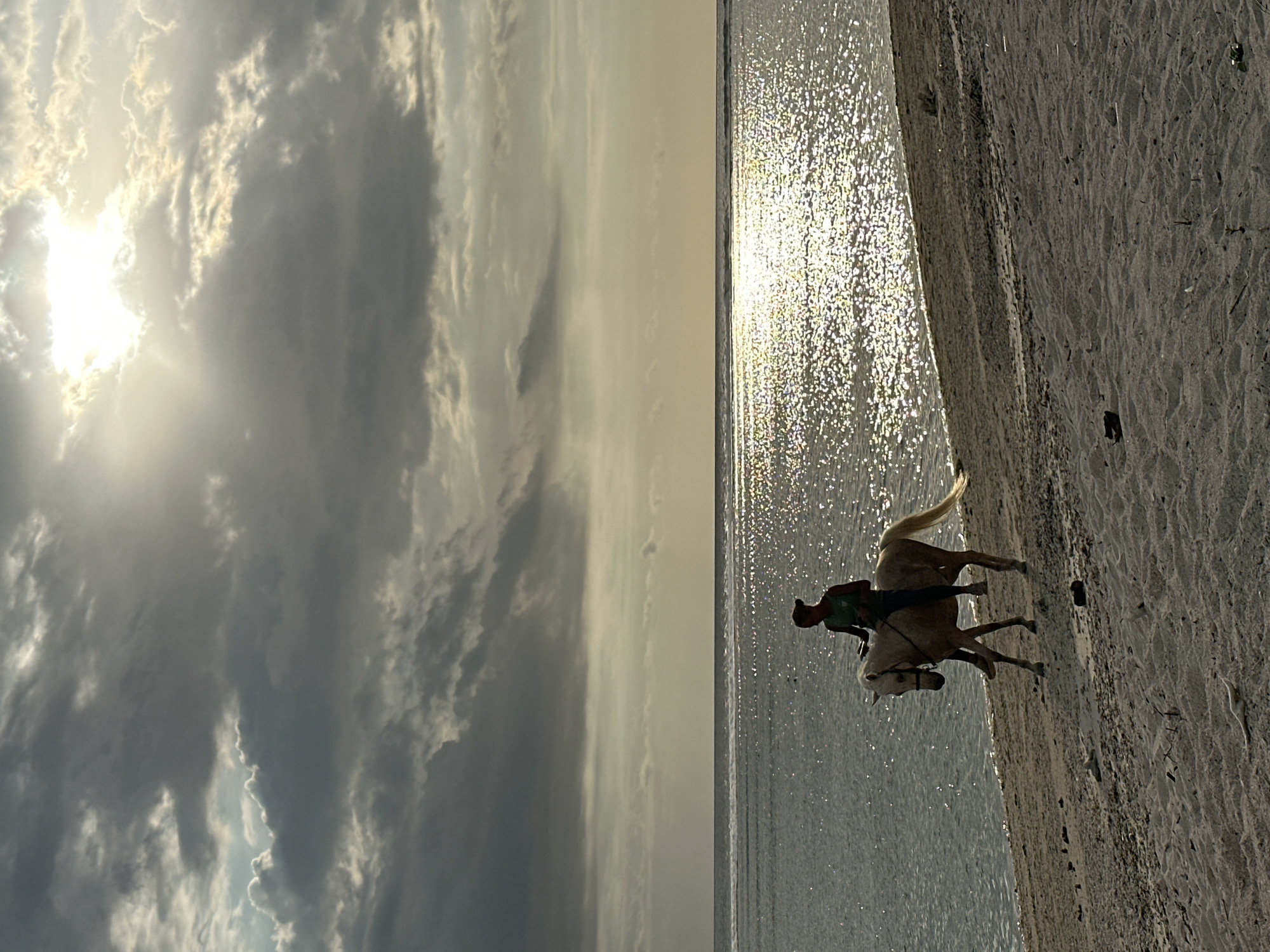 A person rides a horse on a beach in Unknown, Indonesia. The horse is pale with a light-colored tail, and the rider wears dark clothing. The sea reflects sunlight, and clouds fill the sky. The beach has sandy areas.