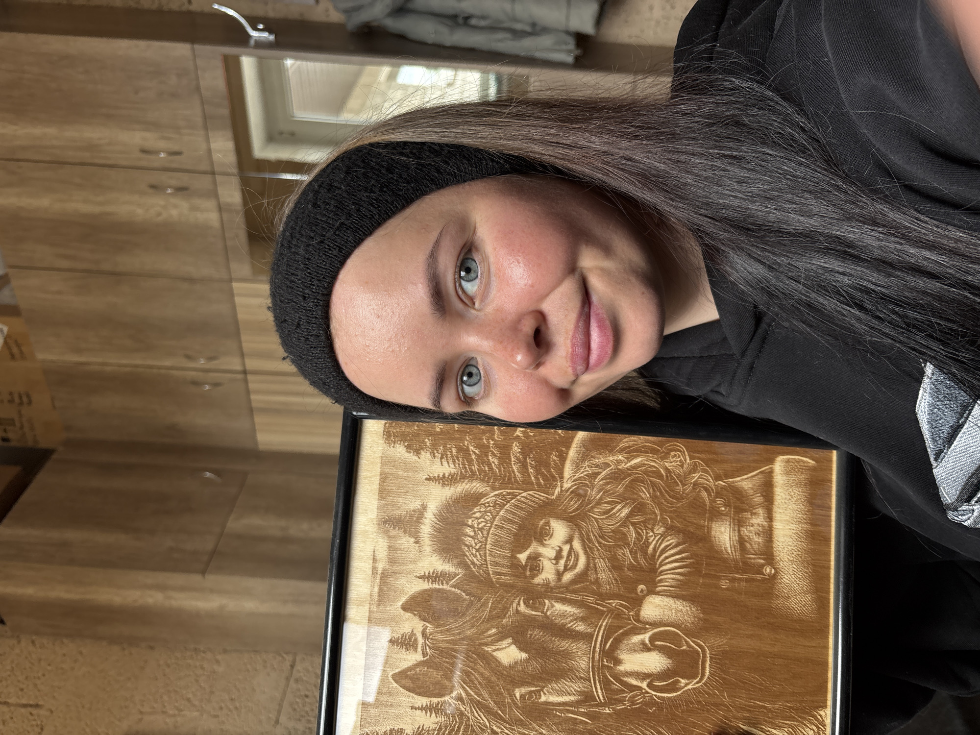 A young woman with long dark brown hair and light blue eyes smiles gently while looking towards the camera, her head tilted. She is wearing a black knitted headband and a black hooded sweatshirt. Her cheeks show a natural flush. She is holding or posing next to a rectangular wooden plaque with an intricate etched or carved image.
The wooden artwork features a depiction of a young girl or woman with long, curly hair, wearing a patterned winter hat, standing beside a horse. The horse's head and neck are visible, and its bridle is detailed. In the background of the carving, tall, conical pine trees suggest a forest or wilderness setting. The artwork is framed in black.
The scene appears to be indoors, likely in a room with warm, wooden cabinetry or paneling. Reflections on a shiny surface in the background show what might be a window or skylight, indicating daylight. The overall atmosphere is personal and engaging, focusing on the woman and the handcrafted artwork. No discernible text is visible on the artwork or in the background. The location context for this image is Русский Вожой, Russia.