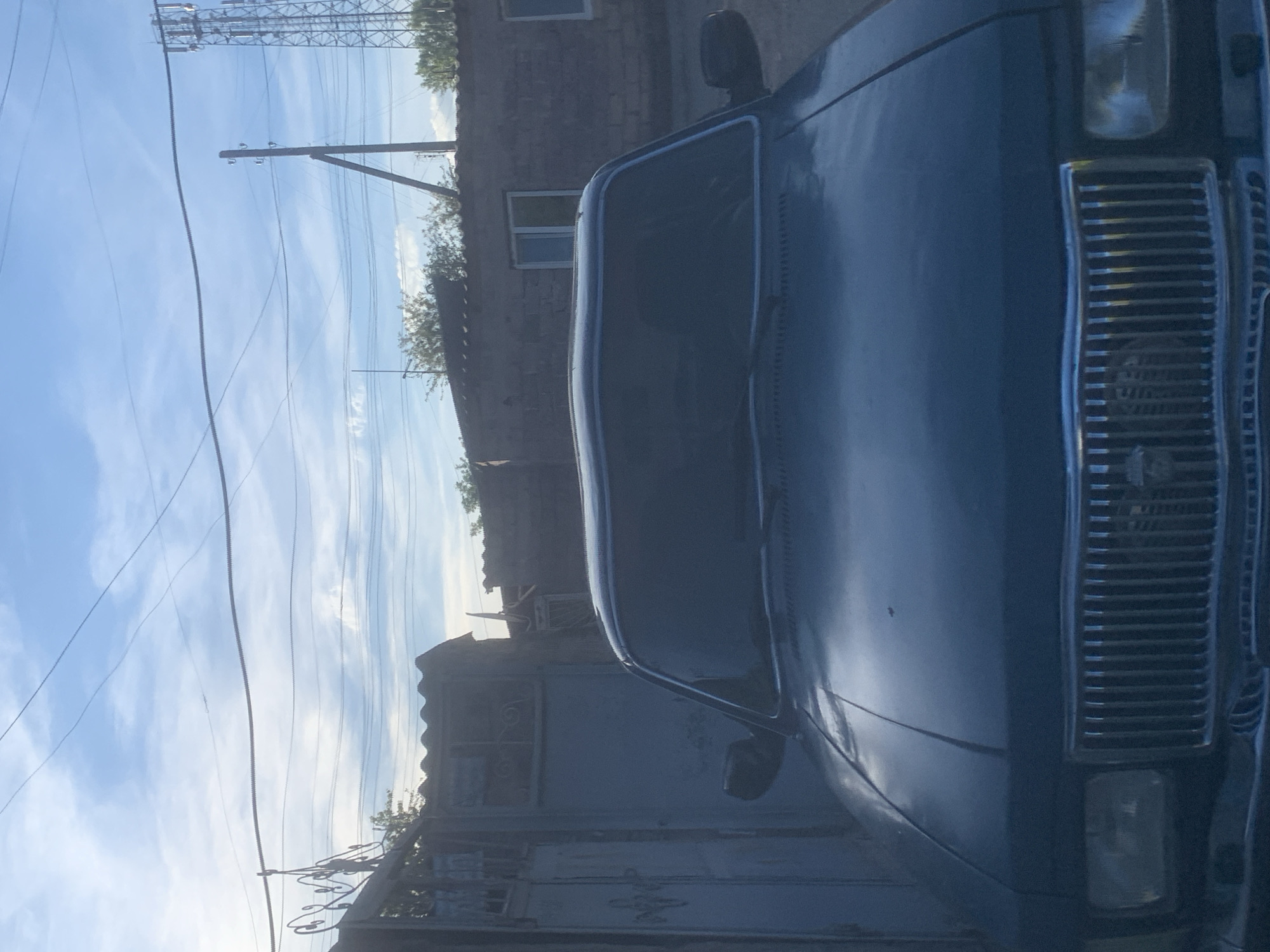 A dark-colored sedan, likely a GAZ Volga based on the vertical chrome-bar grille and the indistinct bird-shaped emblem on the grille, occupies the lower two-thirds of the frame, facing directly towards the viewer. The vehicle features rectangular headlights on either side of the grille and a dark, reflective windshield, obscuring any interior details.
Behind the car, in the midground, multiple structures are visible. To the left, a weathered light-blue metal gate adorned with decorative scrollwork stands next to a building with a dark, possibly corrugated roof. To the right, a brick-textured building features two rectangular windows with white frames and also has a dark roof. Sparse green foliage is visible above and around these structures.
The background consists of a pale blue sky with scattered white, wispy clouds. Numerous overhead power lines, including one appearing thicker and corrugated, traverse the sky horizontally. On the right side, a wooden utility pole, possibly with a supporting strut, stands upright. Further to the right, a section of a tall, metal lattice-structured cellular tower is partially visible against the sky. The scene is represented as being in Gyumri, Armenia; however, the image itself does not contain specific visual identifiers for this location. No individuals or discernible actions are present in the frame.
