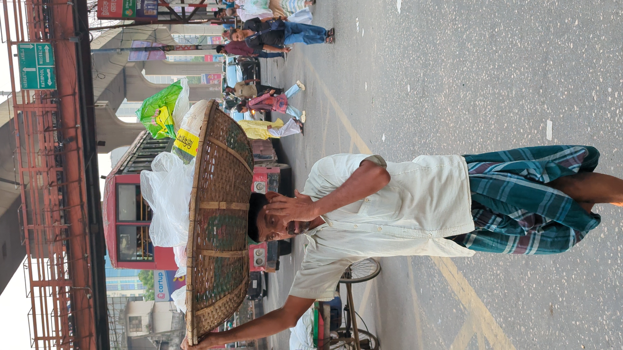 A man is standing on a road in Dhaka, Bangladesh, with a large, round, woven basket balanced on his head. The basket is filled with what appears to be assorted goods, including plastic bags and packages. He is wearing a light-colored shirt and a patterned lungi (traditional South Asian skirt). The man has his hand to his forehead, perhaps adjusting the basket or shielding his eyes.
Behind him, traffic is visible on the road, including a red bus. Above, there is an elevated structure with signs, one of which points towards "HATIRPOOL MO/BAZAR." People can be seen walking on a sidewalk in the background, and there are buildings visible beyond the road. The road appears to be marked with yellow lane lines.