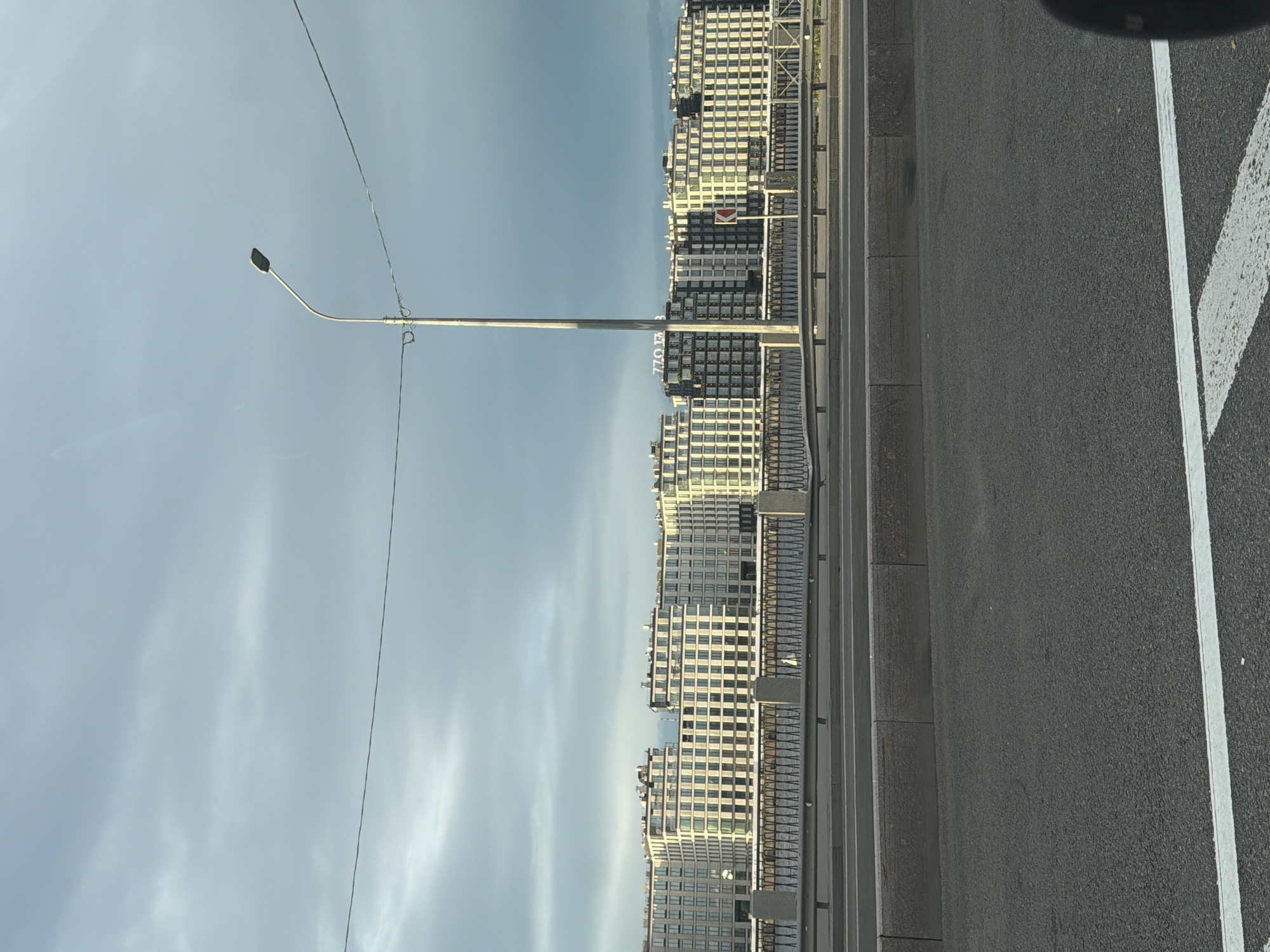 This is a landscape-oriented photograph of a modern city skyline seen from a road. The sky is a pale blue with some wispy clouds, suggesting it could be daytime with possibly overcast conditions. On the left side of the frame, a tall, slender street lamp with a curved arm extends into the sky, with a thin wire running from it towards the right. The dominant feature is a row of tall, multi-story buildings that stretch across the middle ground. These buildings have a uniform, grid-like facade with many windows, suggesting residential or office complexes. Some have balconies or terraces.
The foreground consists of an asphalt road with white lane markings, indicating a street or highway. To the left of the road, there is a concrete curb and a metal railing, separating the road from the buildings. The perspective implies the photo was taken from a vehicle moving along the road, or from a vantage point alongside it.
There is some visible text on one of the buildings. It appears to be "770 173", possibly a street number or building designation. There is also a small red and white sign on one of the buildings that resembles a traffic sign. The overall mood is neutral, focusing on the urban architecture and infrastructure. The time of day appears to be either early morning or late afternoon due to the soft lighting.