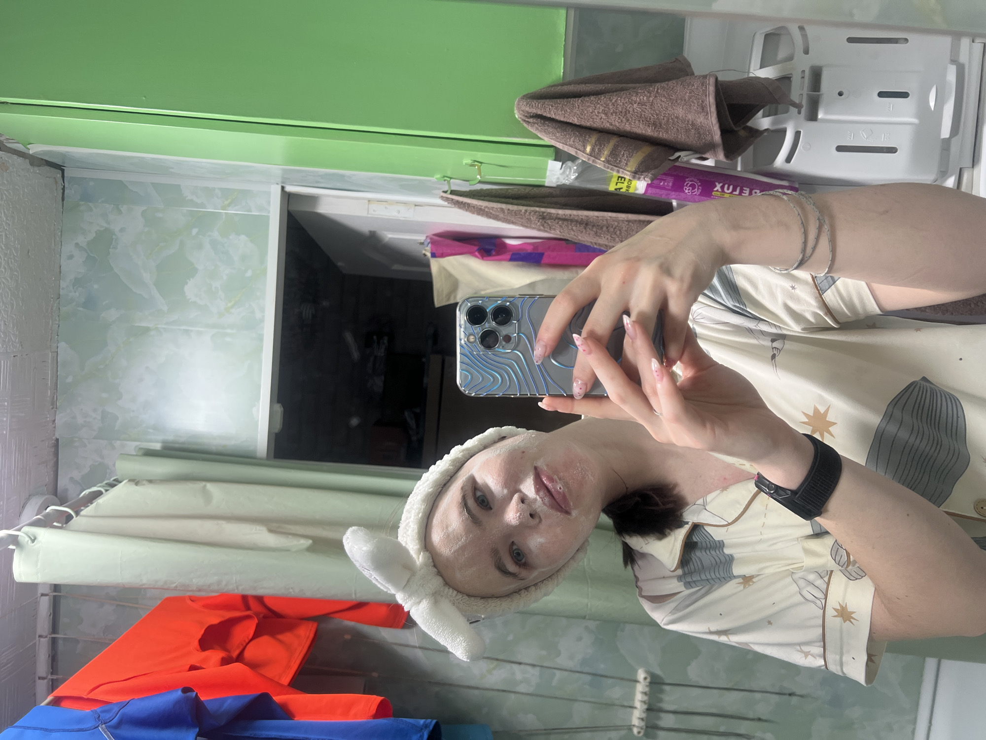 A mirror selfie captures a person engaged in a self-care routine within what appears to be a bathroom. The subject, a female with dark hair pulled back, wears a white fluffy headband with bunny ears. Her face is covered with a white skincare product or mask, with visible product residue around her mouth and nose. She is holding a light blue iPhone with a distinctive wavy-patterned case, using it to take the photo in the mirror. Her nails are manicured with light-colored polish and small speckles.
She is wearing light-colored pajamas or loungewear, featuring a pattern of grey shapes and small brown stars. A black smartwatch or fitness tracker is visible on her left wrist, along with a few thin, light-colored bracelets on her right arm.
The background, reflected in the mirror, shows typical bathroom elements. To the left, there's a light-colored shower curtain with faint horizontal stripes. Below it, a drying rack holds vibrant orange and blue shirts. To the right, behind the person, a green wall or cabinet is visible above a white storage unit or shelf containing two brown towels and a purple bottle. The bottle shows inverted Cyrillic text, part of which reads "ЕЛВ" and "БЕТОХ / DETOX," consistent with the provided location context of Chaikovsky, Russia. The ceiling area is a solid light green, and parts of the walls feature light green and white marbled tiles.