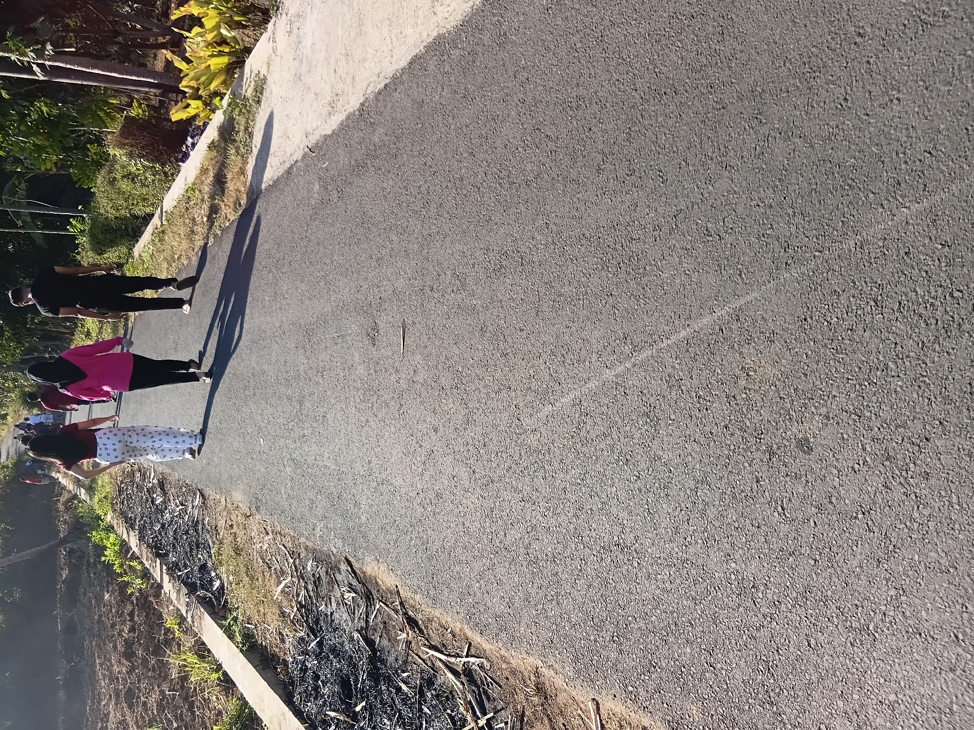 Three individuals are walking away from the camera on a paved road in Tanggul Wetan, Indonesia. The person in the center wears a pink headscarf and a pink top, while the person to their left wears white pants with a black pattern and a dark red top. The person to the right wears a black t-shirt and black pants. The road is bordered by a dry, barren strip of land with scattered debris on the left and vegetation on the right. Trees are visible in the background. The lighting suggests it is daytime, likely morning or afternoon, with shadows cast on the road.